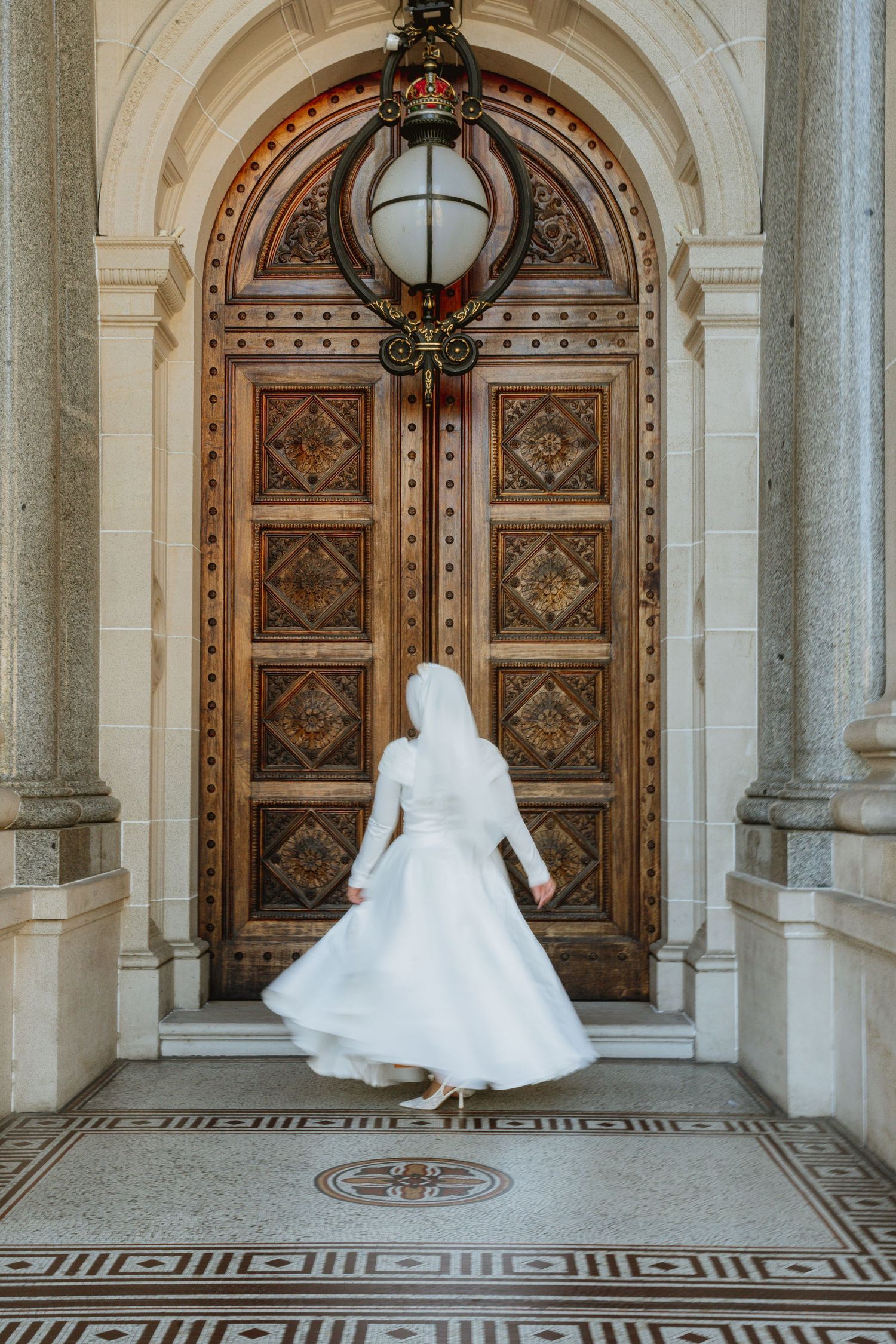 Bride in white wedding dress and hijab standing before ornate wooden doors of elegant stone building.