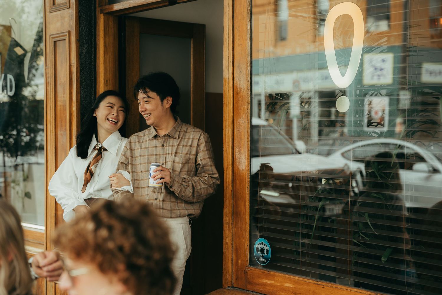 Two smiling people standing in a cafe doorway holding coffee cups on a sunny urban street.