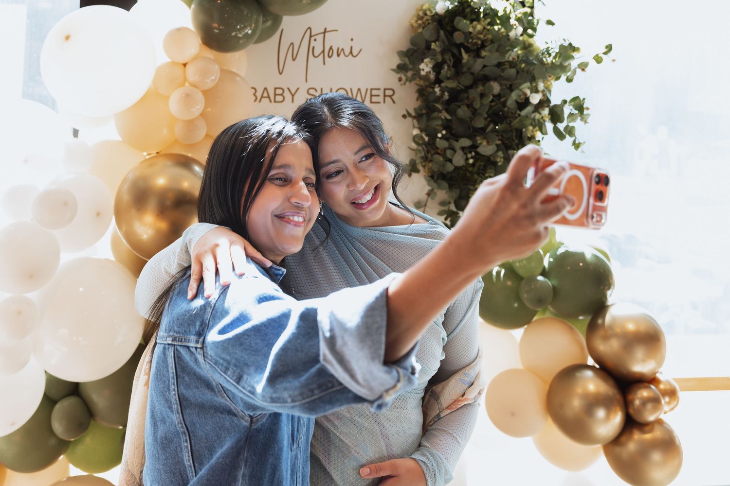 Two people take a selfie together while embracing, surrounded by gold and white balloon decorations.