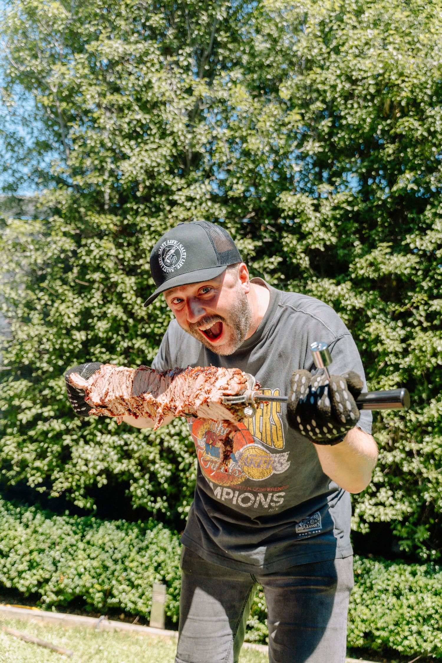 Person holding up a large smoked or grilled meat outdoors with trees in background.