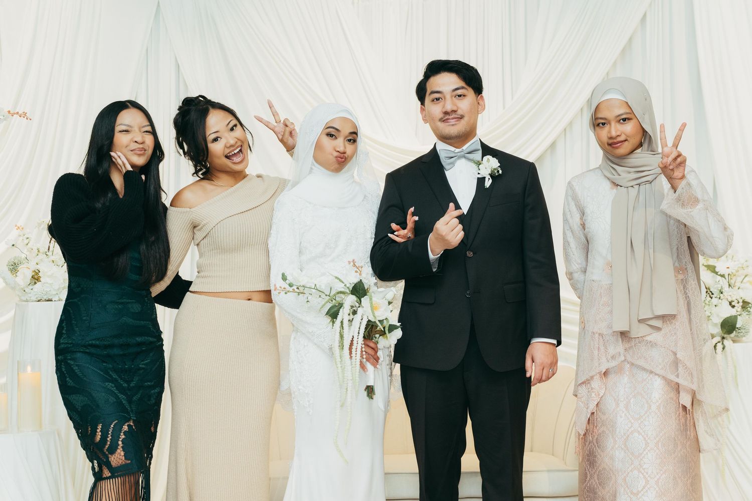 Muslim bride and groom posing with friends making peace signs at wedding reception.