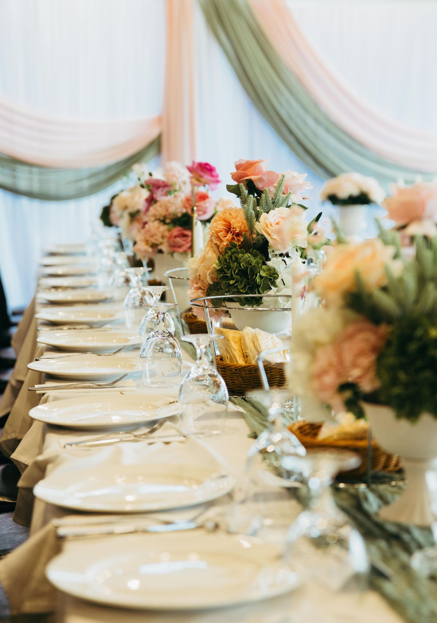 Elegant wedding reception table with floral centerpieces, white plates, and draped fabric backdrop.