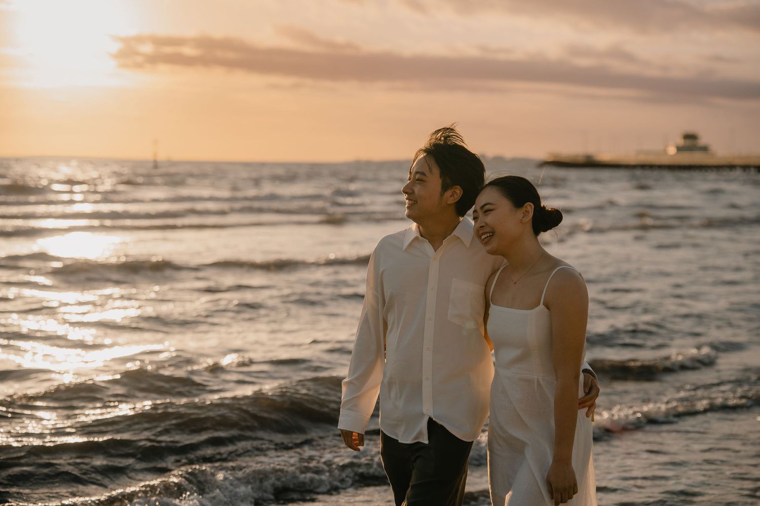 Couple walking hand in hand along beach at golden sunset, woman in white dress, man in white shirt.