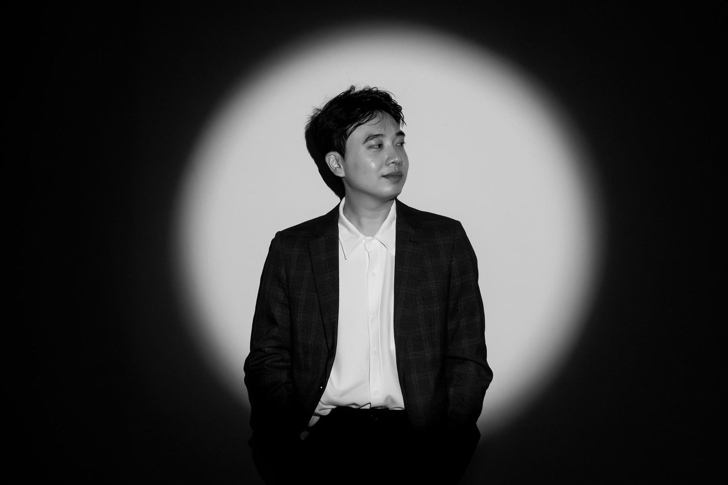 Young Asian man in dark blazer poses under spotlight in black and white portrait photography.