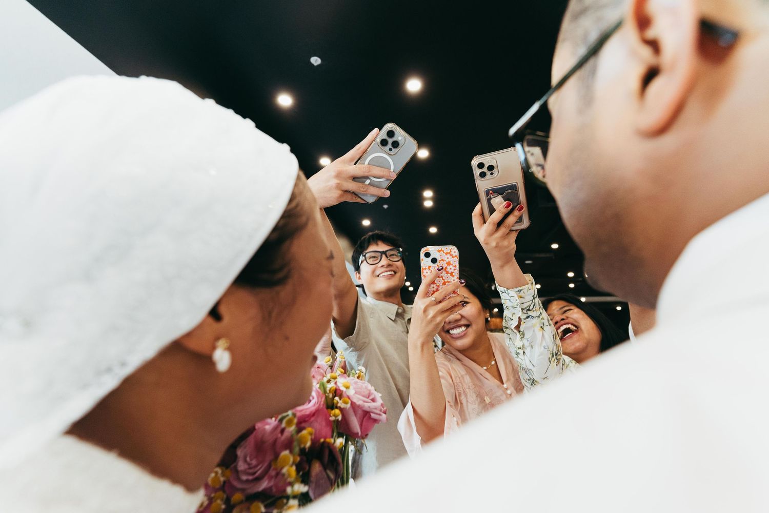 Wedding guests photograph smiling bride and groom with smartphones at joyful reception.