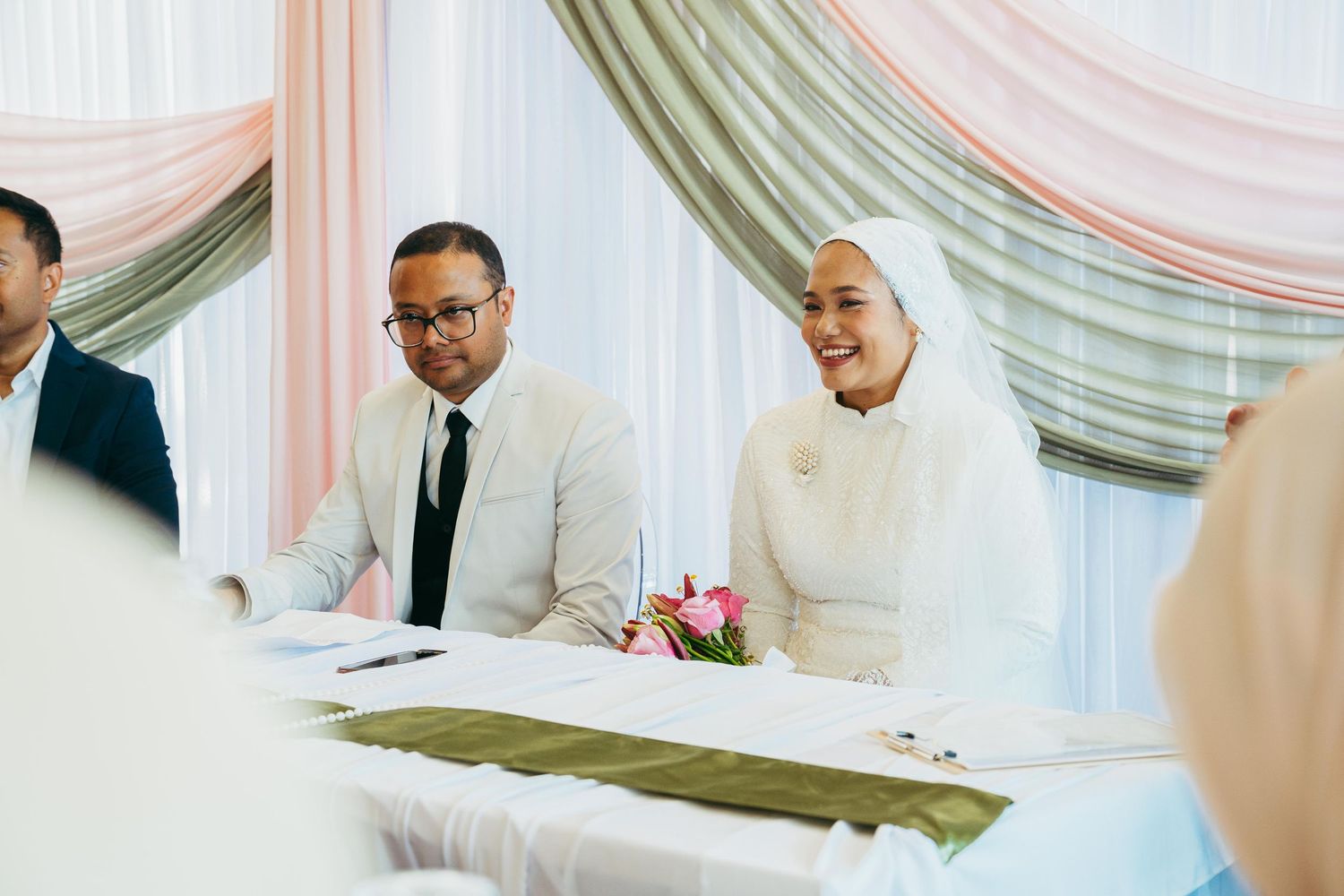 Muslim couple smiling at wedding ceremony table, bride in white hijab holding pink flowers, elegant draped backdrop.