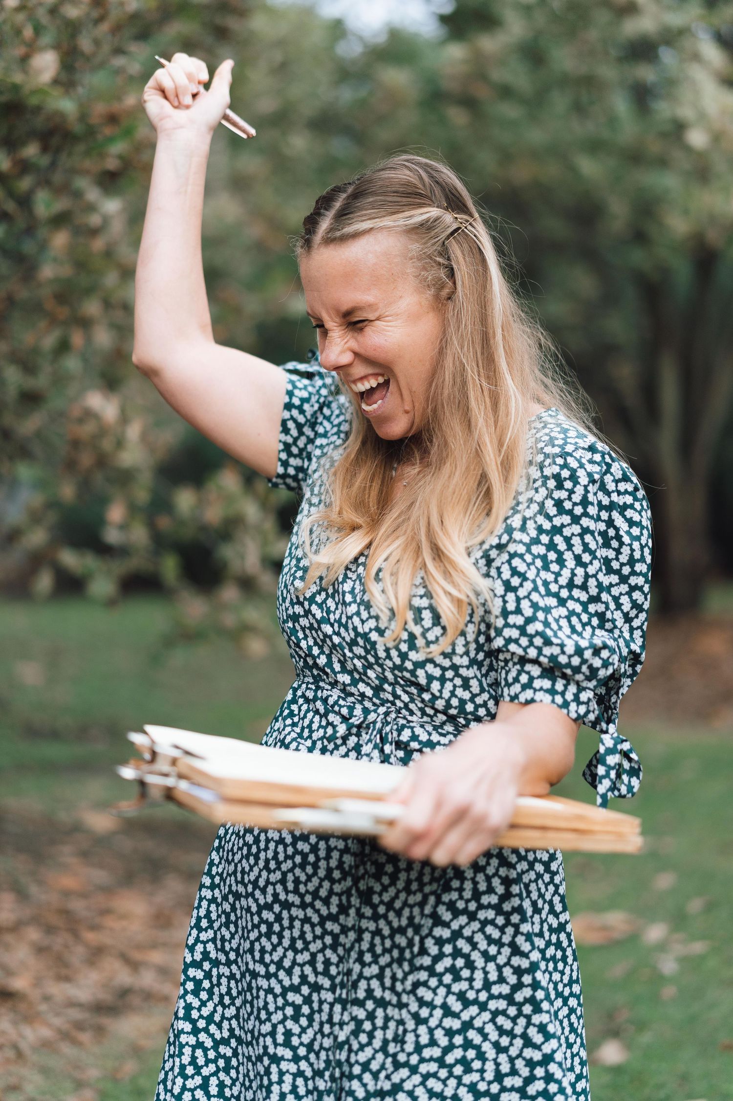 Excited blonde woman in floral dress holding wooden frames and raising hand outdoors.