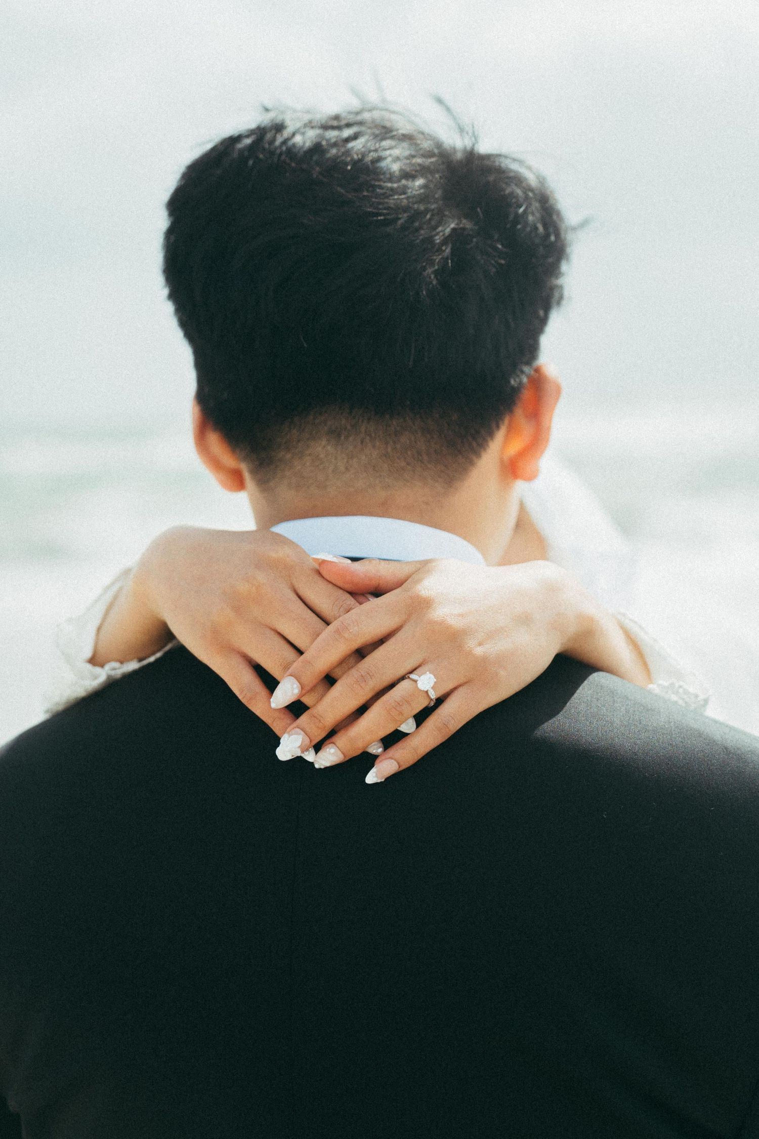 Bride embracing groom from behind showing diamond engagement ring and white manicured nails at beach wedding.