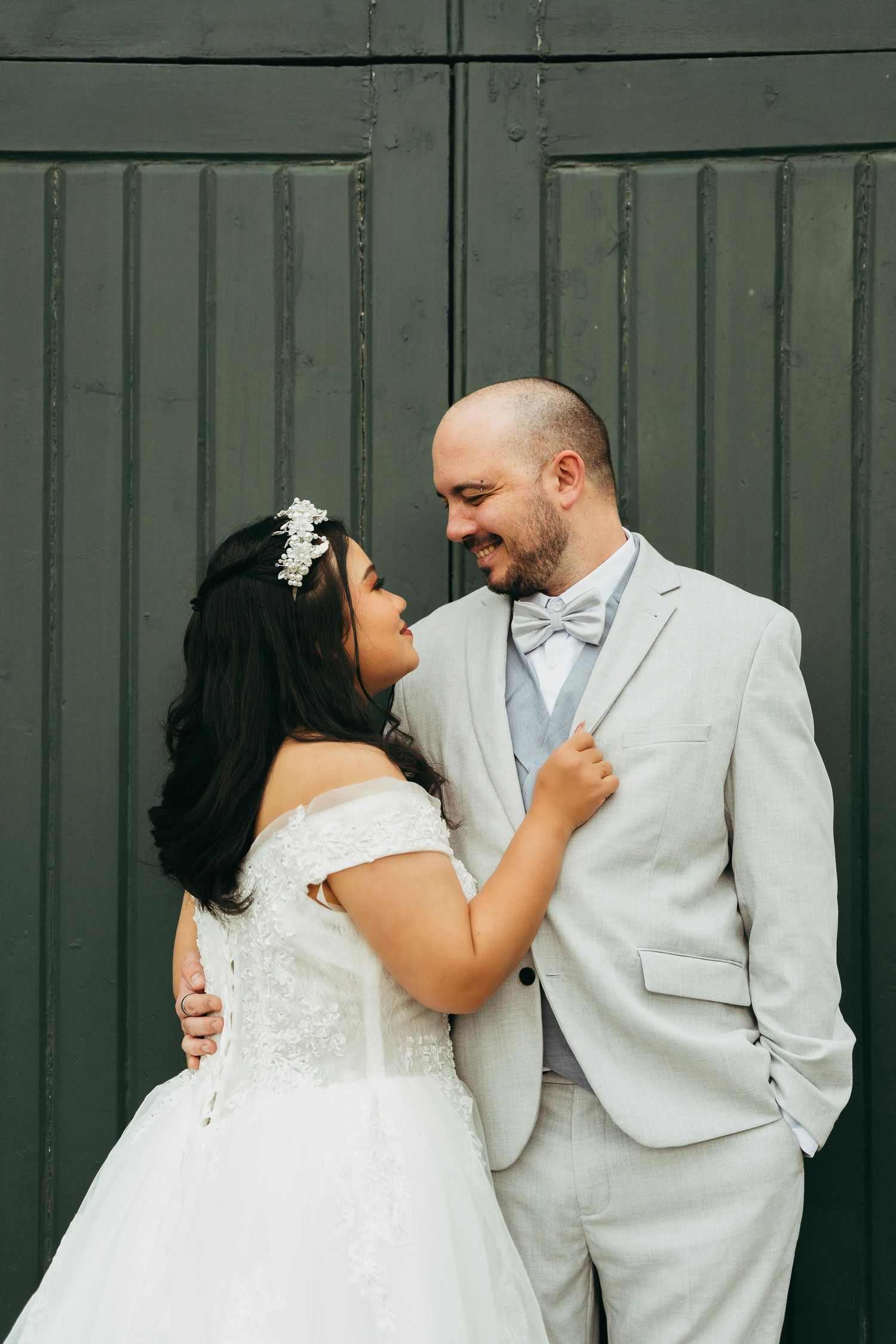 Bride and groom share a romantic moment in front of a dark green door, wearing a white gown and gray suit.