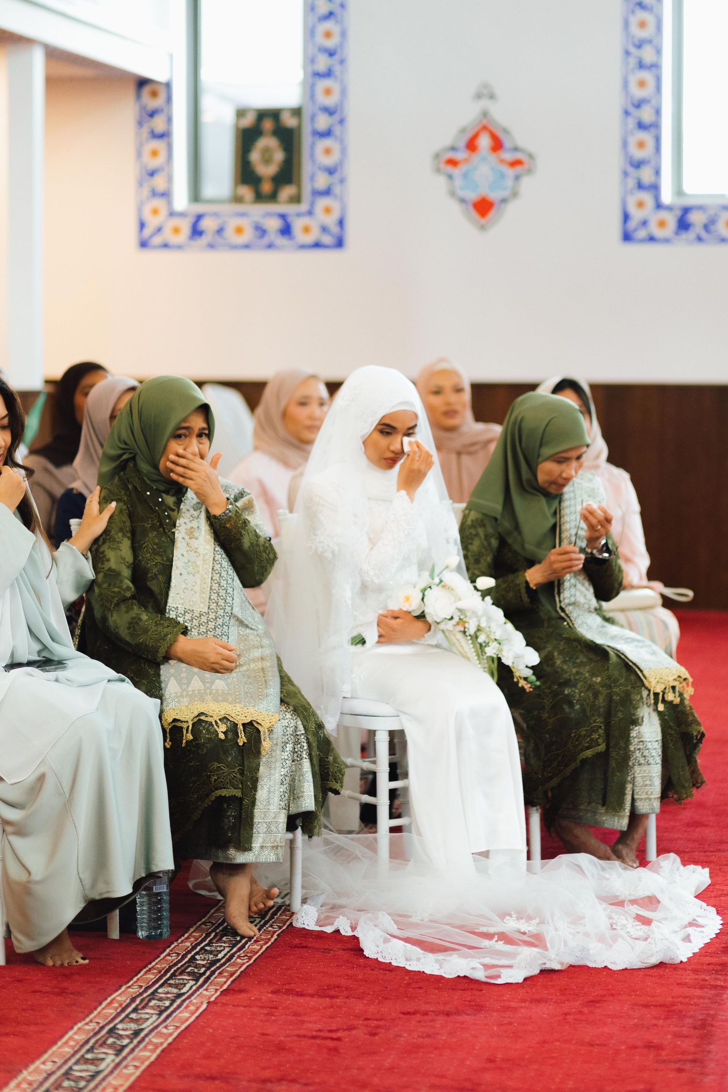 Emotional Muslim bride in white hijab and gown crying during Islamic wedding ceremony surrounded by guests in green hijabs.