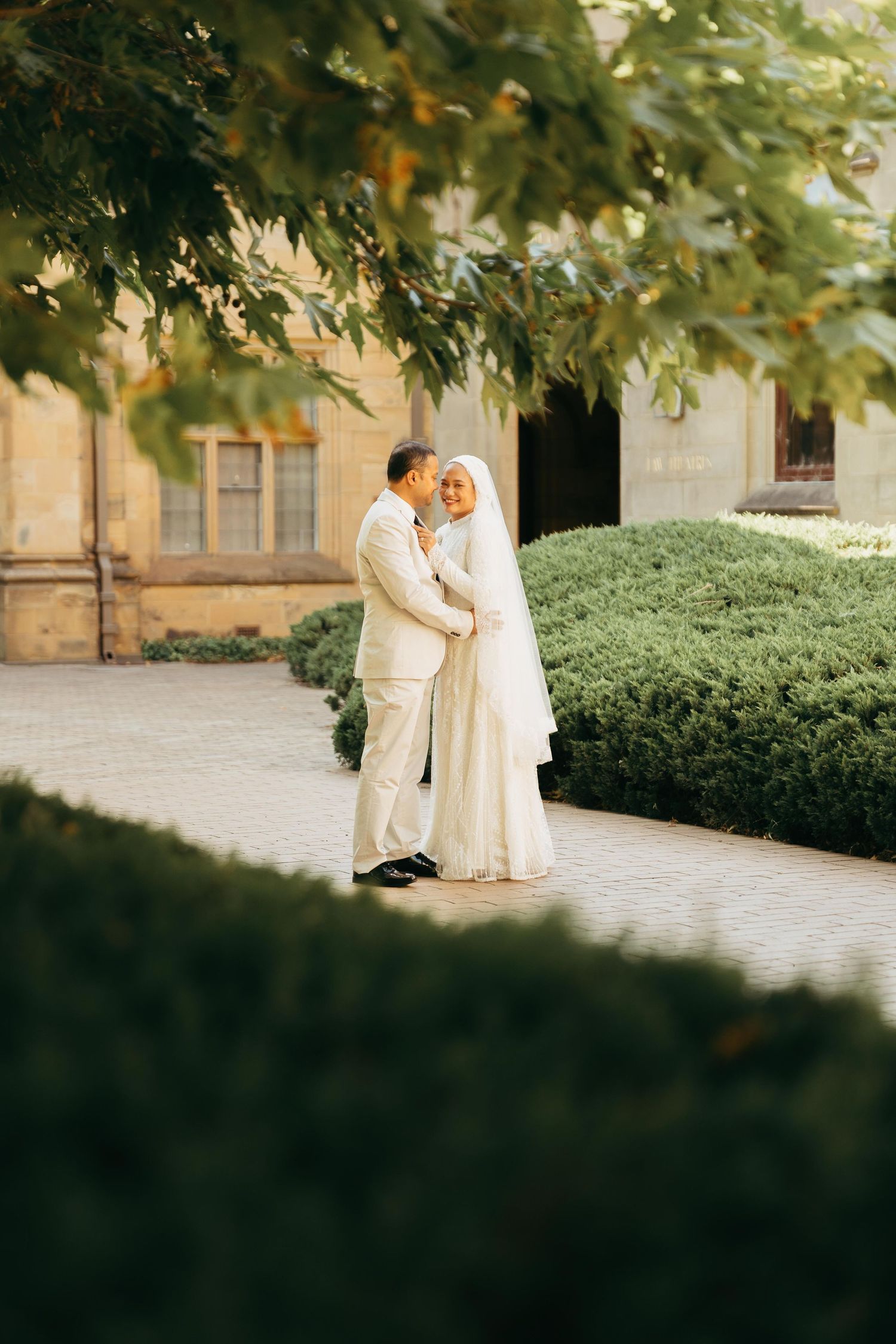 Muslim bride and groom share intimate moment in lush garden courtyard of historic stone building.