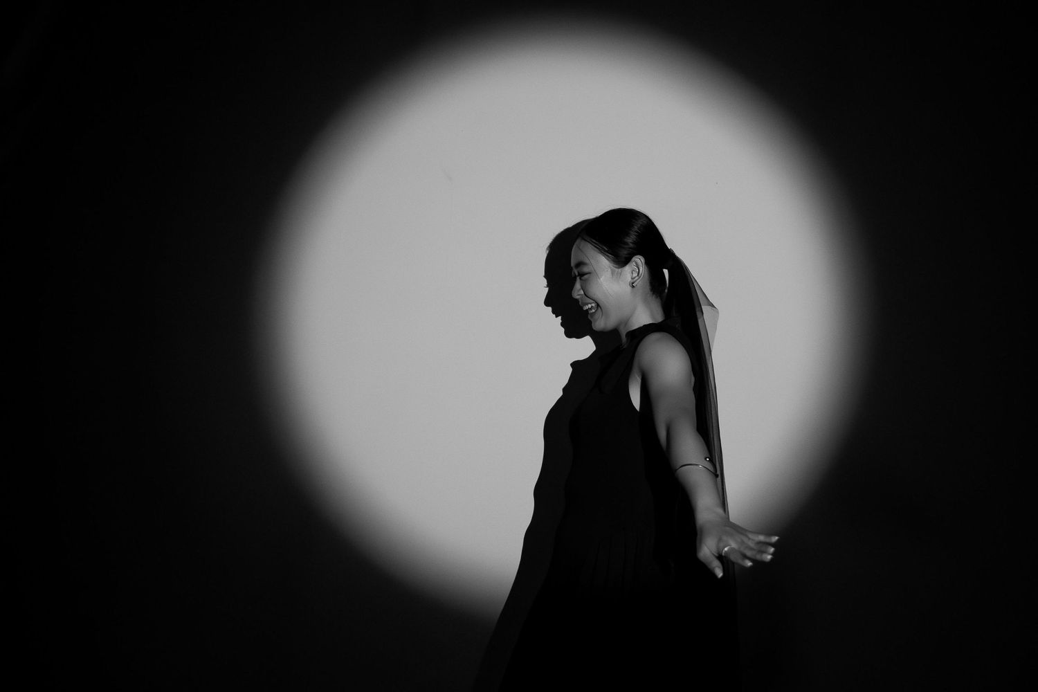 Black and white silhouette of a woman in a sleeveless dress standing in a circular spotlight on dark background.