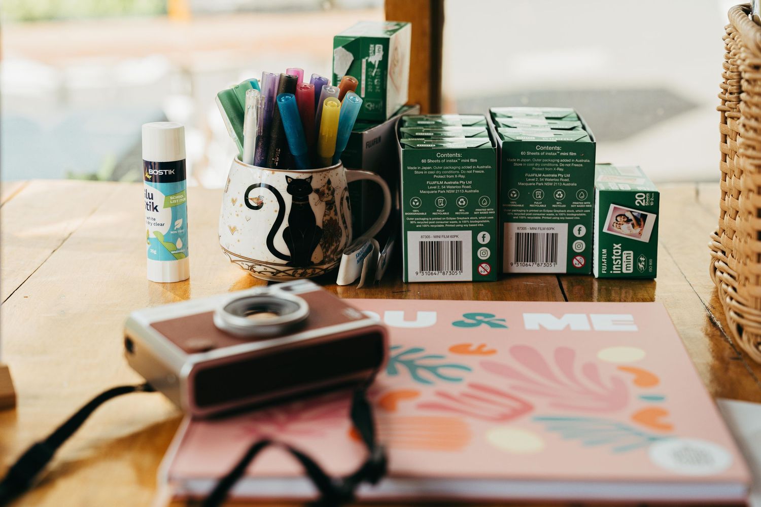 Wedding guest book table with Instax camera, colorful markers, film boxes, and floral notebook on wood surface.