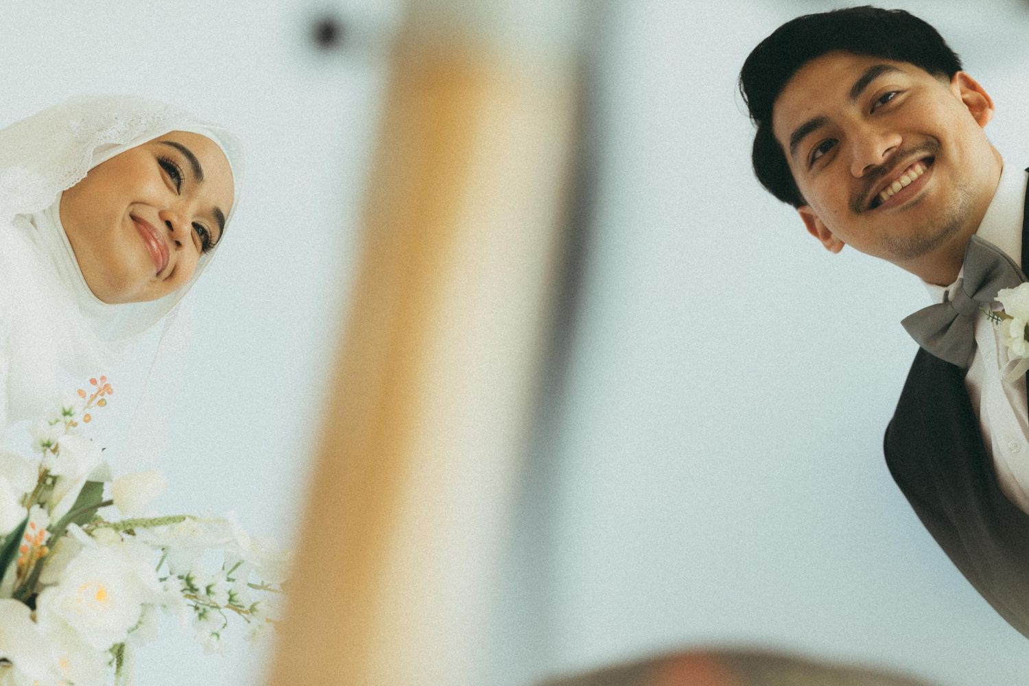 Happy Muslim bride and groom smiling up at camera from below holding wedding bouquet of white flowers.