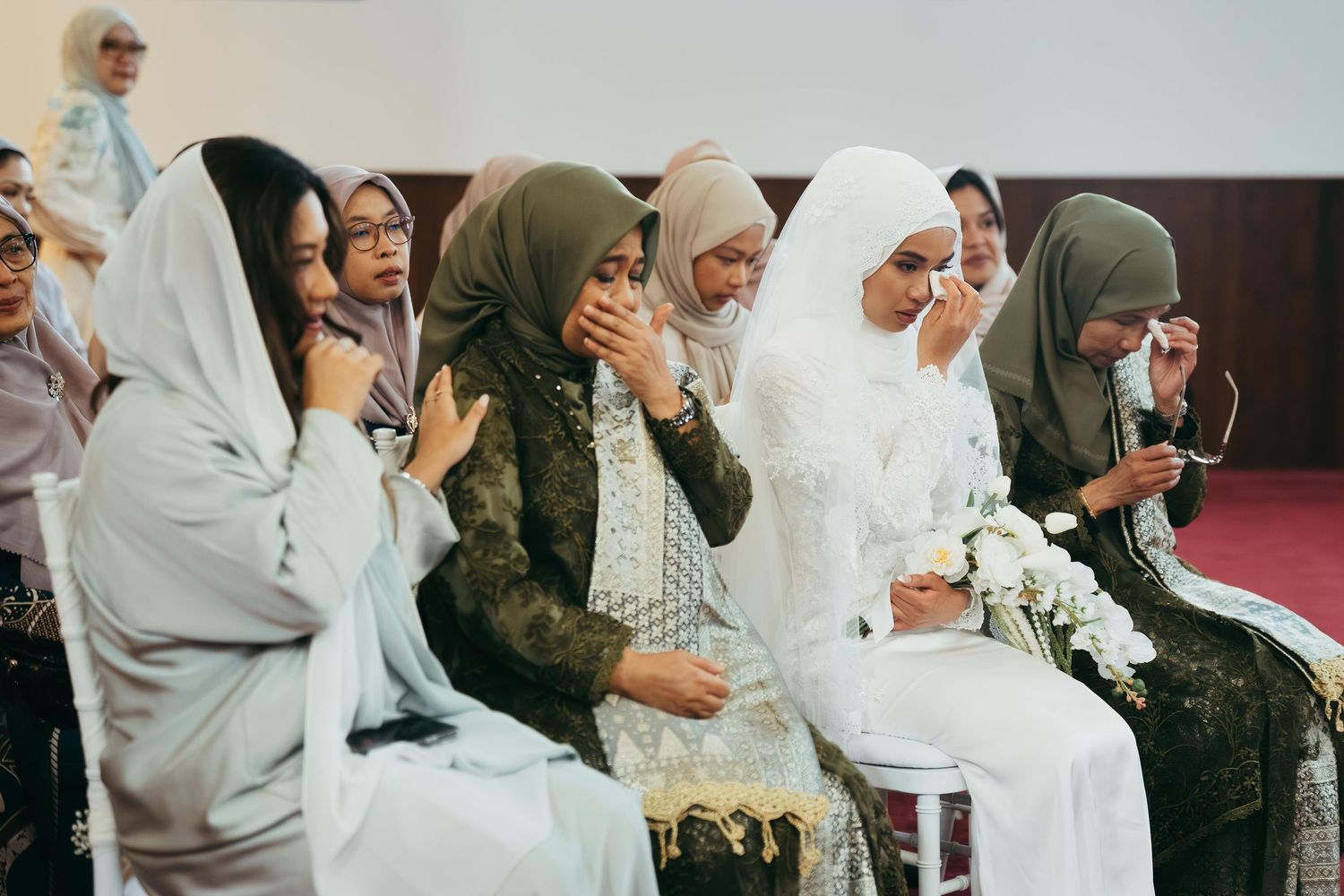 Muslim women in hijabs crying emotionally during a wedding ceremony, seated in rows at a formal indoor venue.