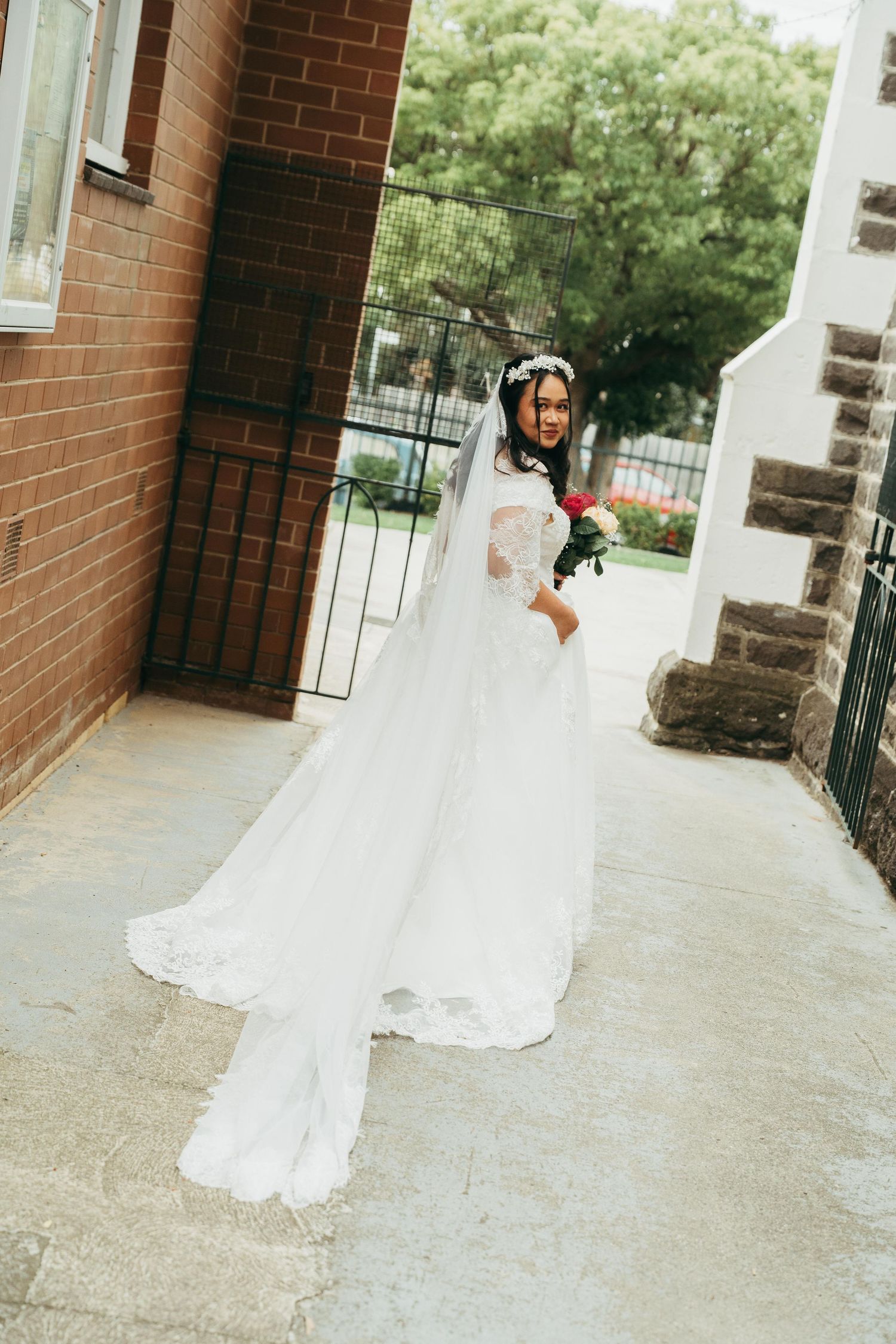 Elegant bride in a white lace gown and veil holds a bouquet while posing on church steps outdoors.