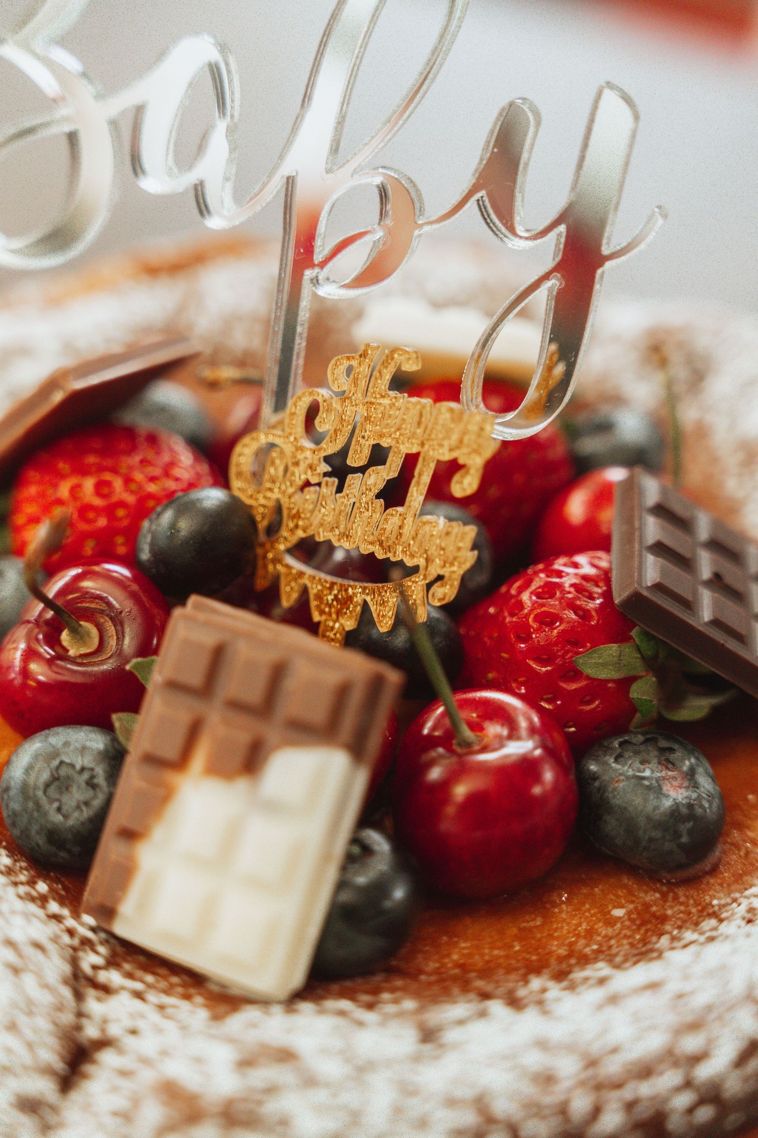 Close-up of chocolate cake decoration with fresh strawberries, blueberries and gold birthday topper on powdered sugar surface.