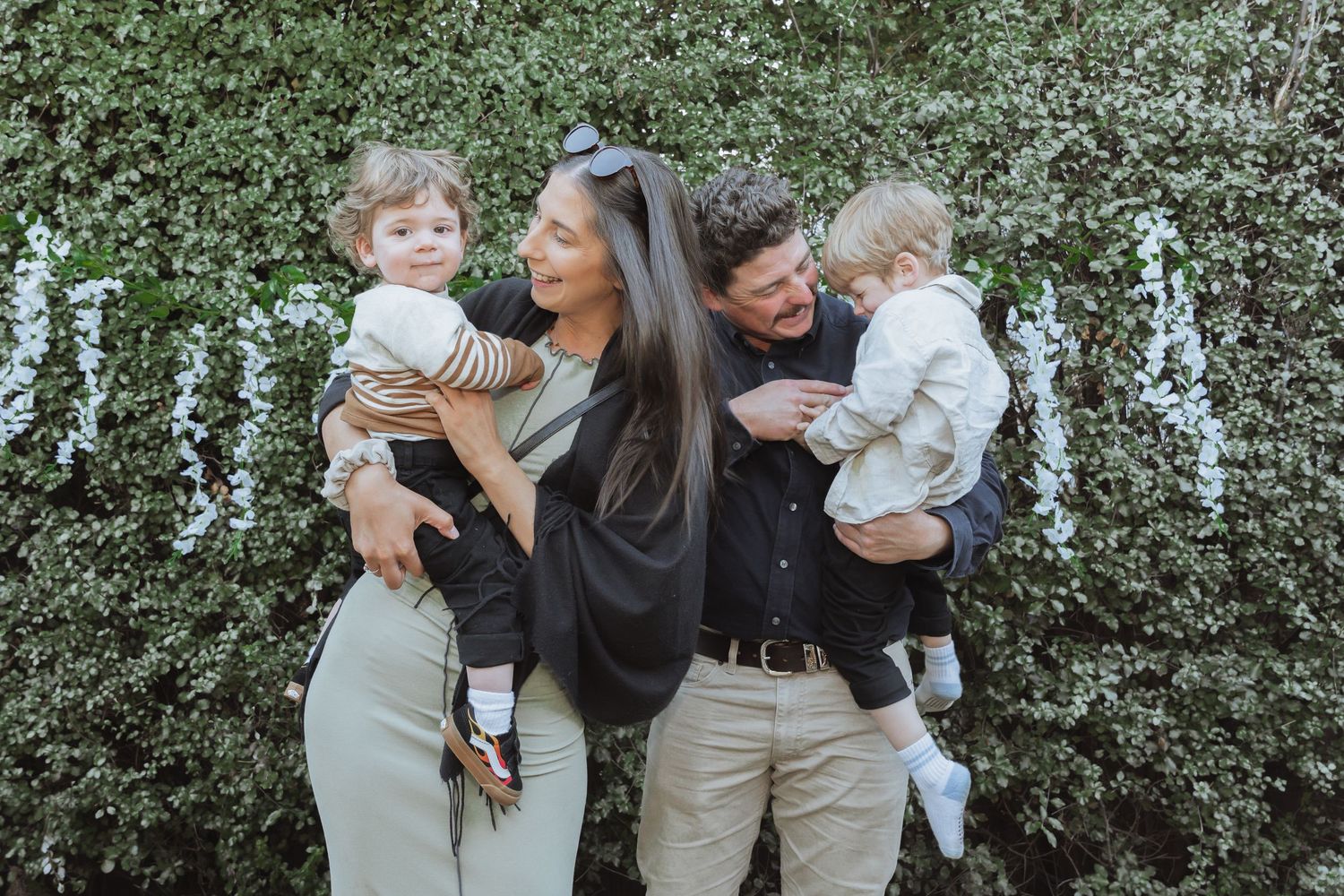 A family of four poses together in front of a flowering bush with white blooms.