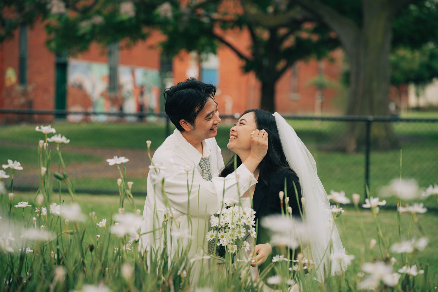 A couple in wedding attire shares a romantic moment among white wildflowers in a lush green park setting.