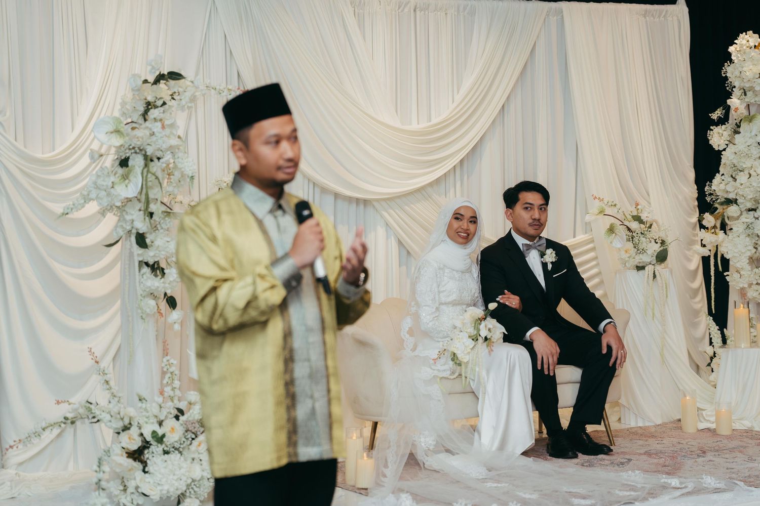 A man in a gold traditional outfit speaks at a Muslim wedding ceremony while the bride and groom sit on stage.