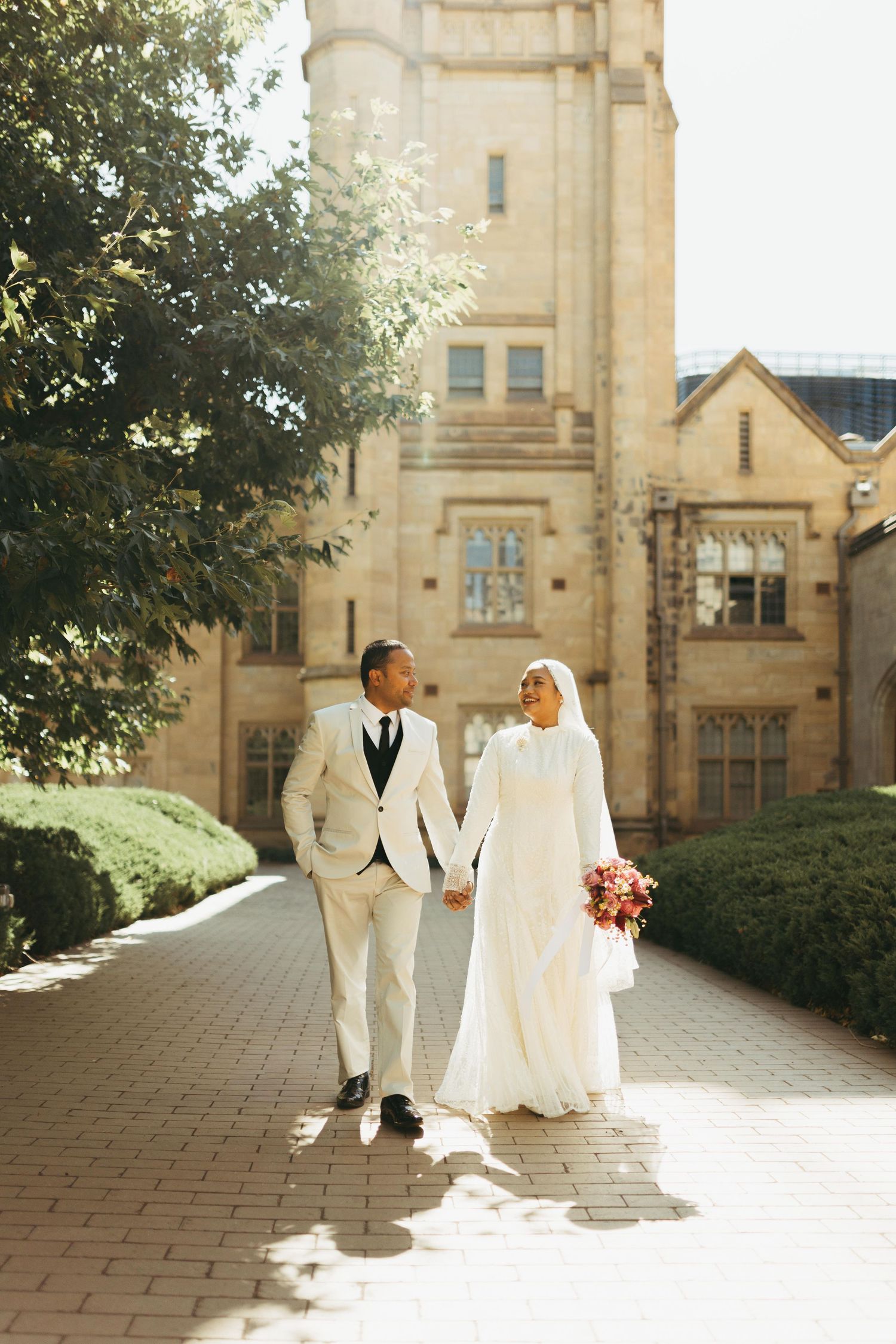 Muslim newlyweds walk hand in hand along sunlit courtyard path before grand historic stone building.