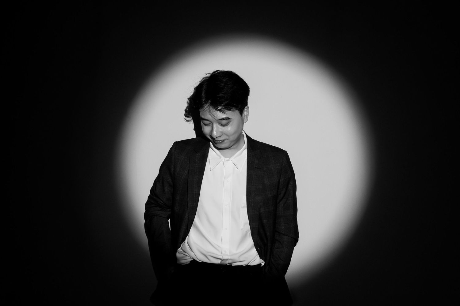 Black and white portrait of a man in a blazer standing under a spotlight, looking downward thoughtfully.