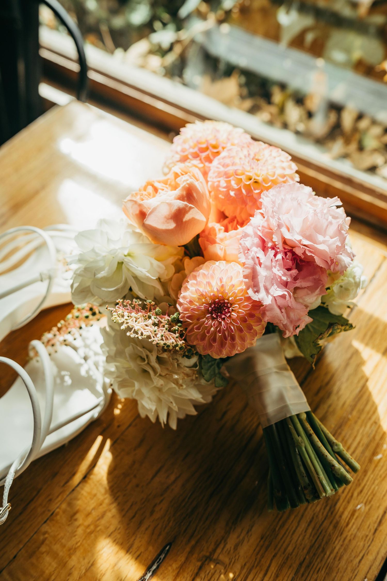 Bridal bouquet of peach, pink, and white flowers with ribbon, beside white heels on a wooden table in sunlight.