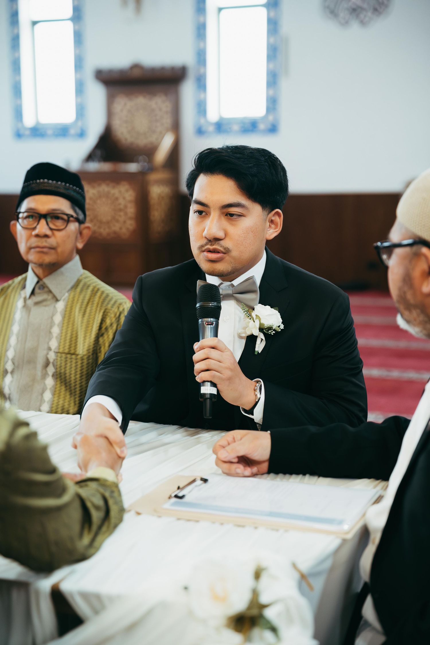 A groom in a black tuxedo speaks into a microphone during an Islamic wedding ceremony at a mosque.