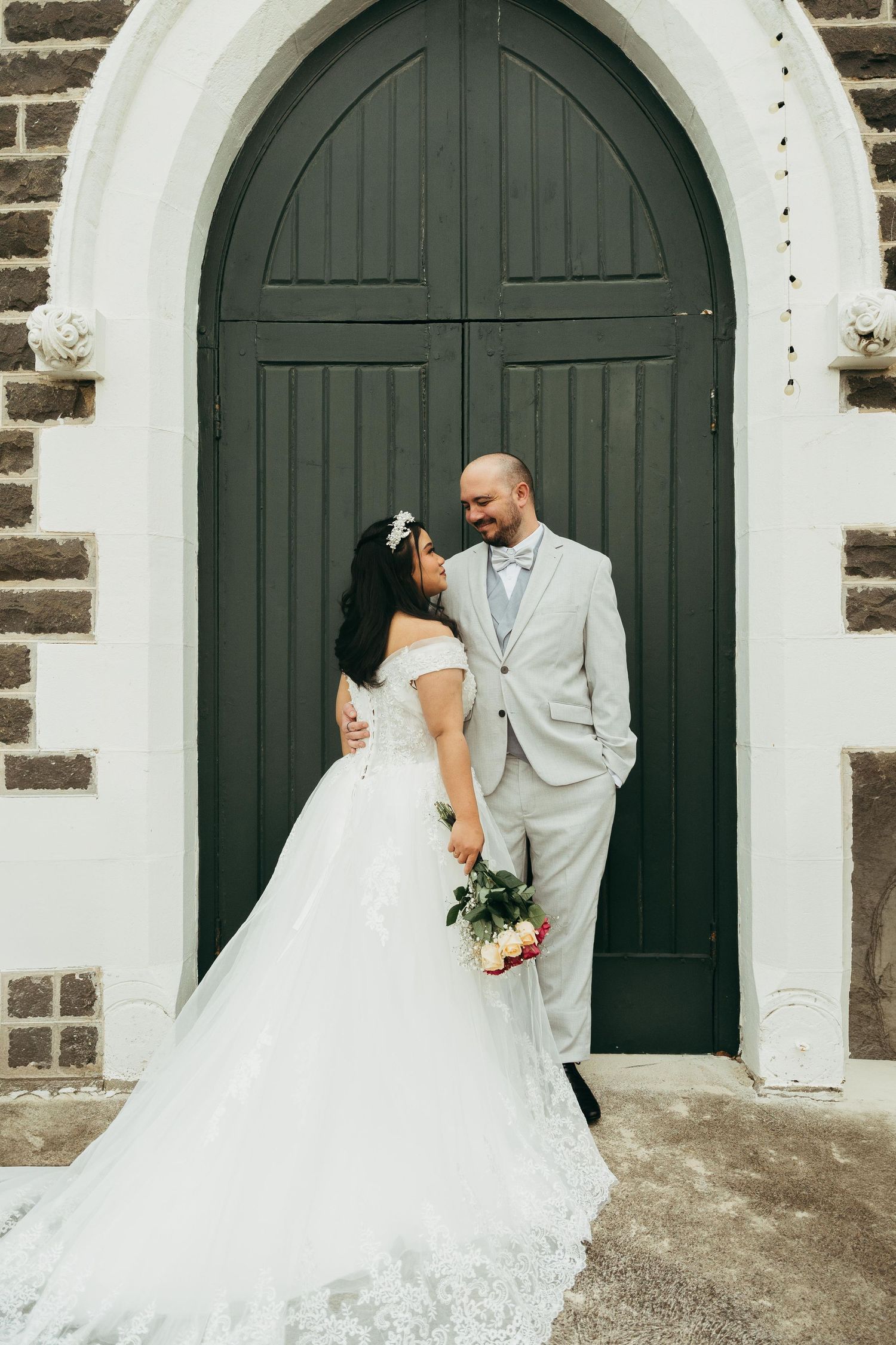 Smiling bride and groom pose together holding a bouquet before a large arched green wooden church door.