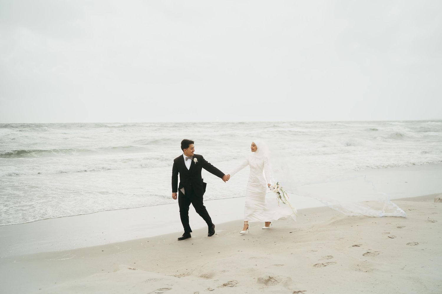 A bride in a white hijab and groom in a black suit walk hand in hand along a sandy beach.