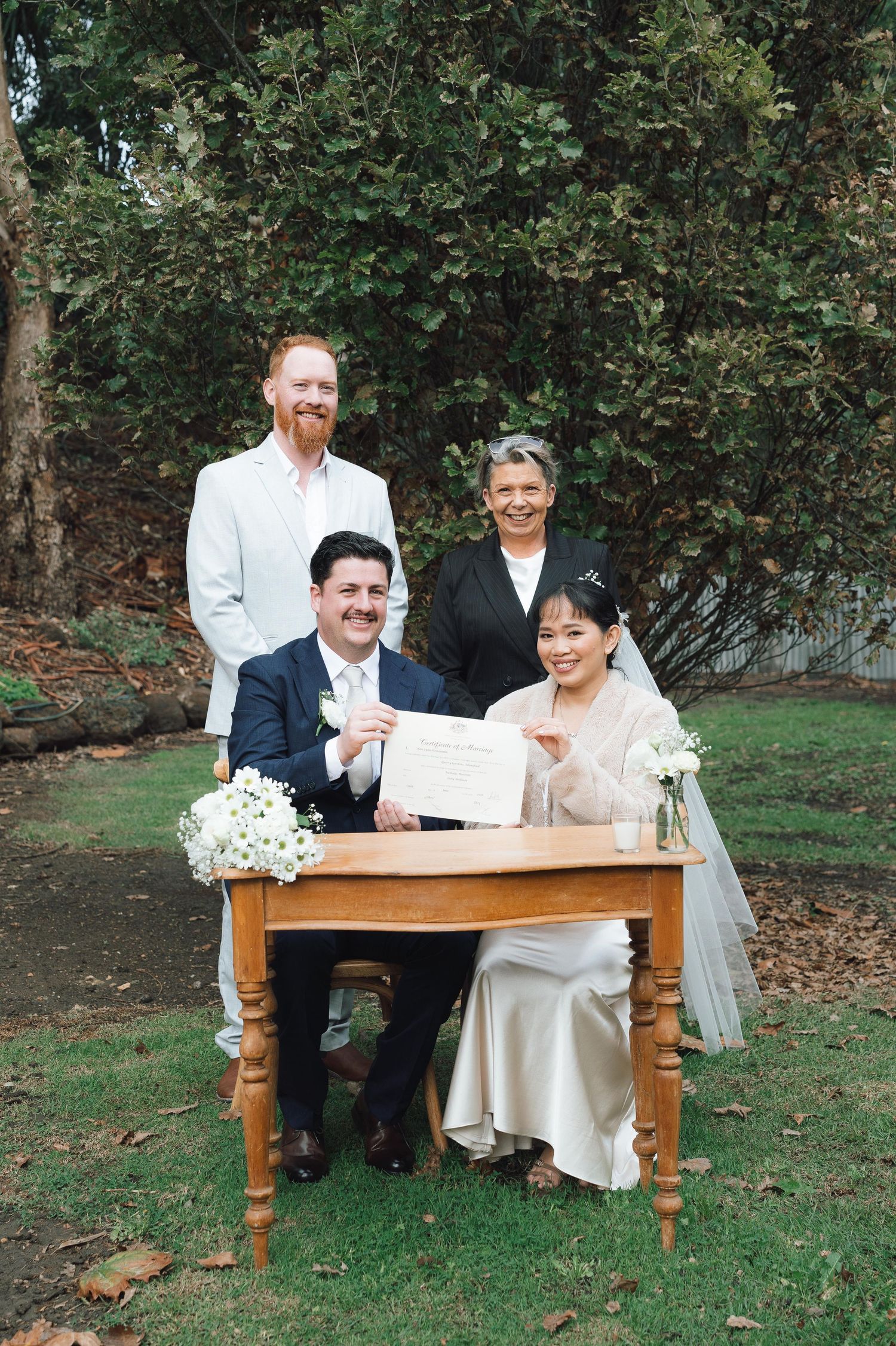 Bride and groom sign wedding certificate at wooden table outdoors, joined by two witnesses in a lush garden setting.