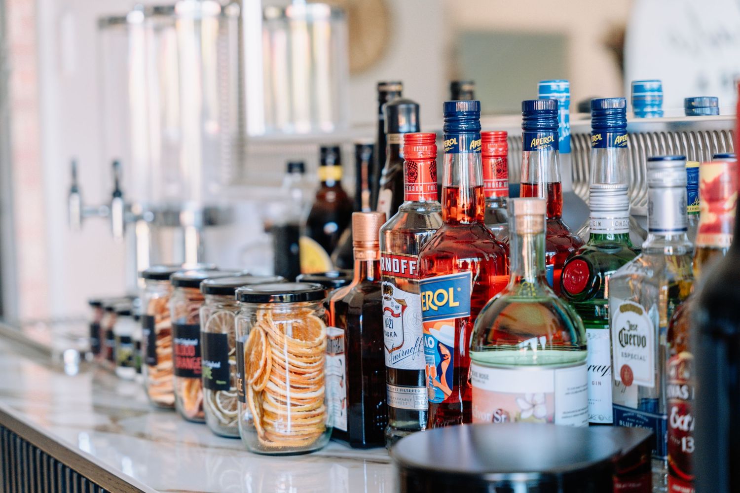 Various liquor bottles and mixers arranged on a bar counter with glass jars and cocktail supplies.