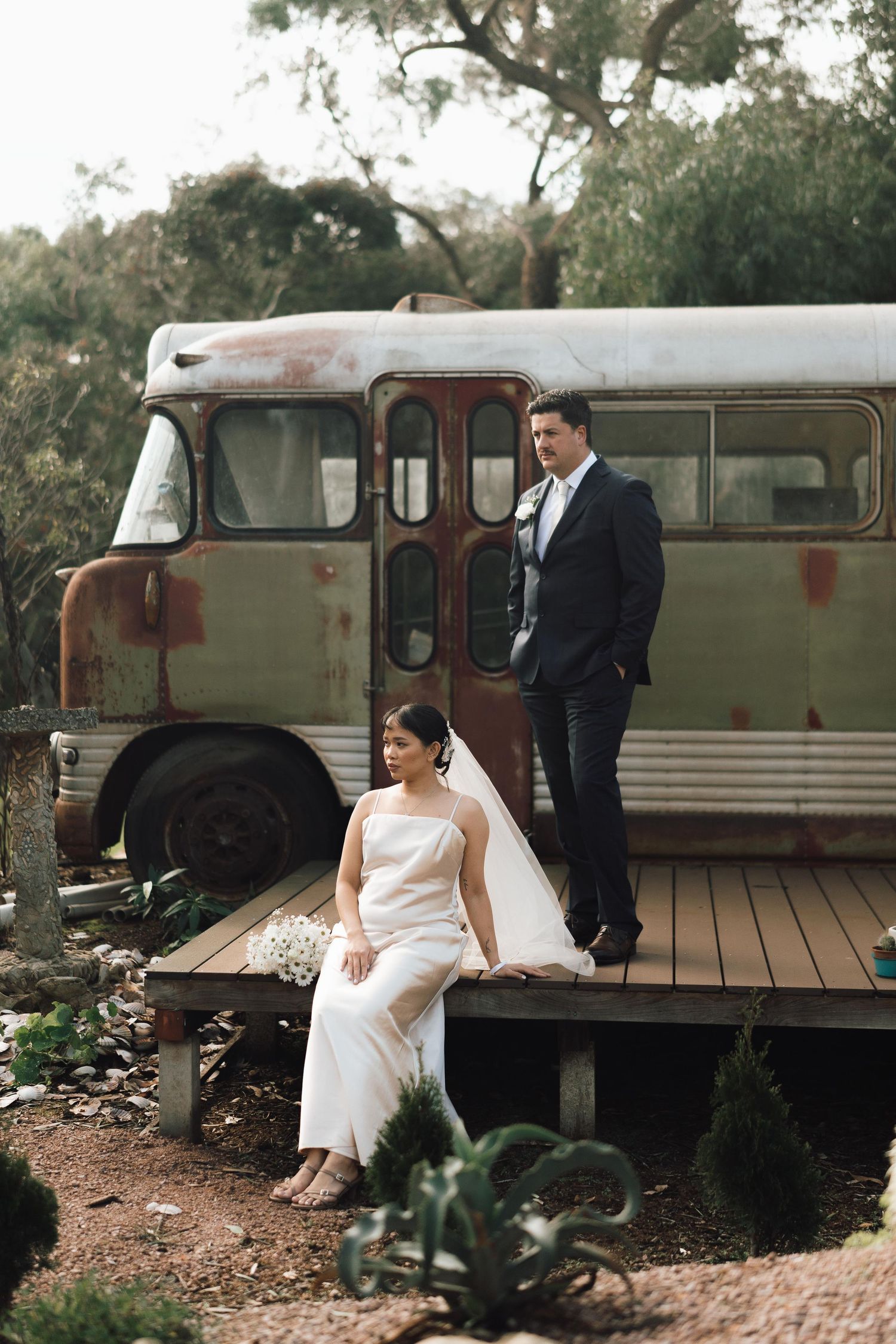 Bride and groom pose by rustic vintage bus on wooden deck in garden setting.