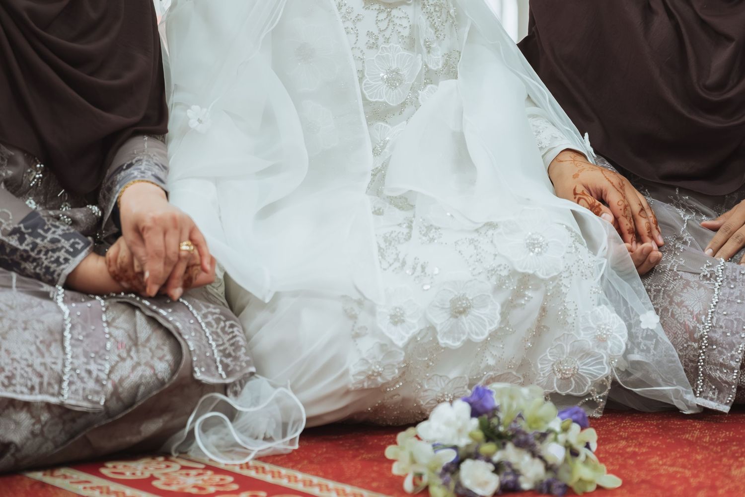 Bride and groom in traditional Islamic wedding attire holding hands with a bouquet nearby.