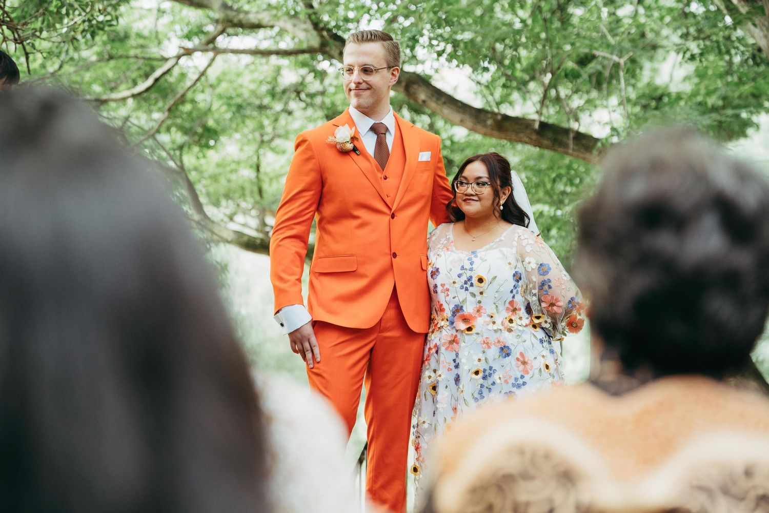 Groom in bold orange suit walks down outdoor wedding aisle with smiling woman in floral dress.