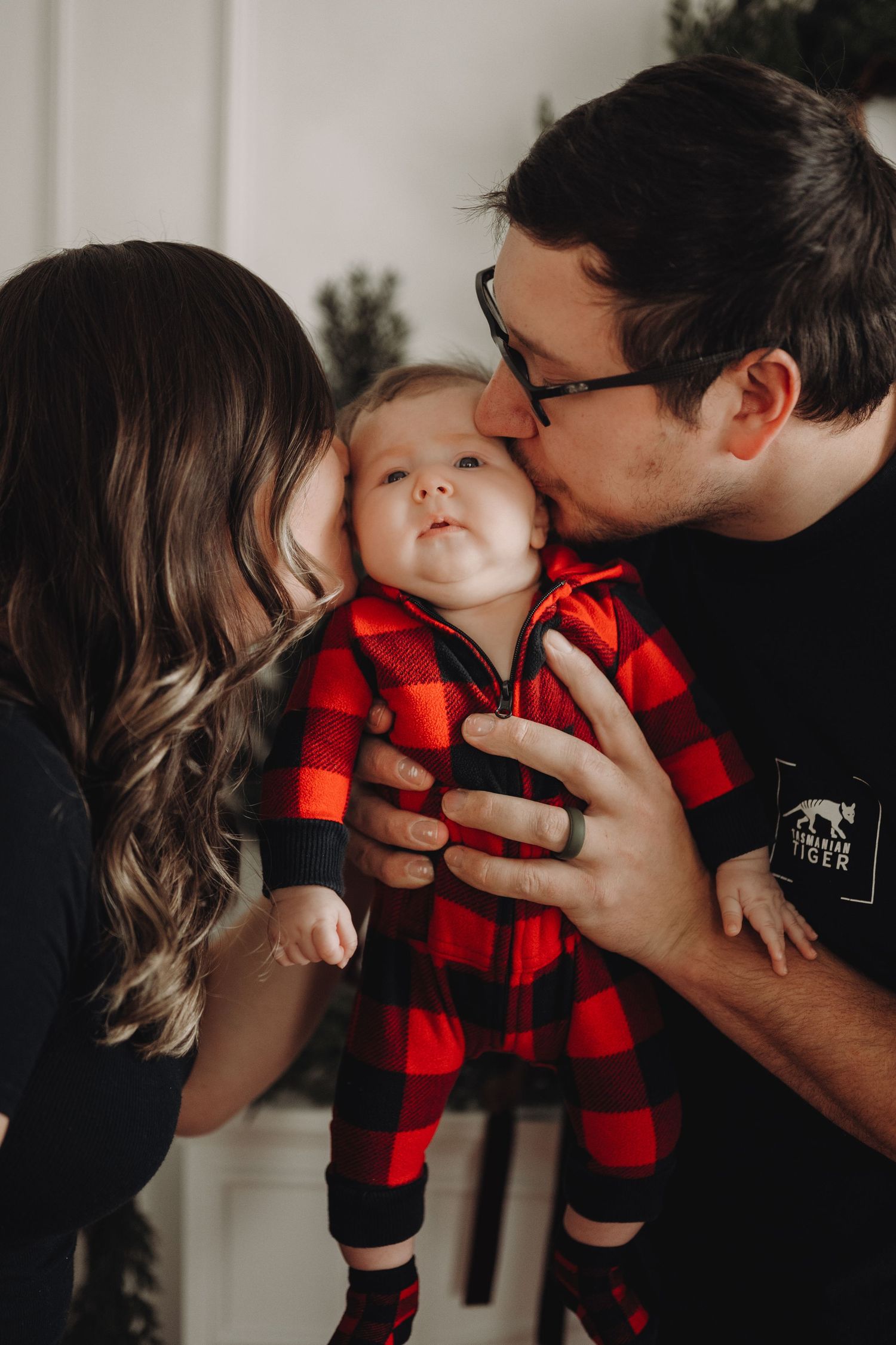 Parents kissing their baby in red plaid pajamas during a cozy Christmas family photo session.
