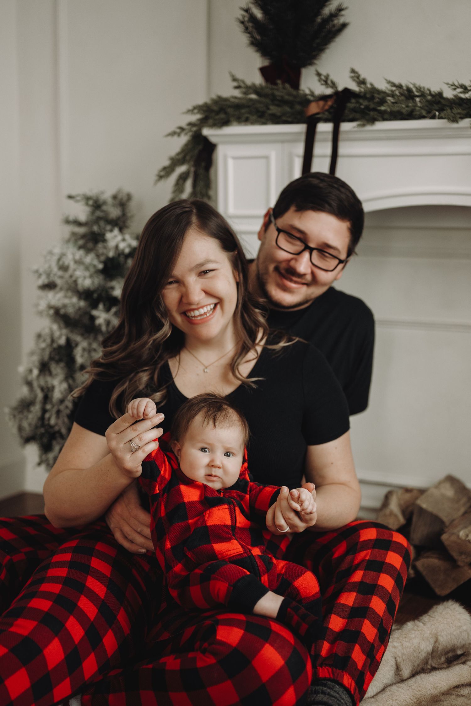 Family in matching red plaid pajamas sitting by fireplace with Christmas tree, smiling parents holding baby.
