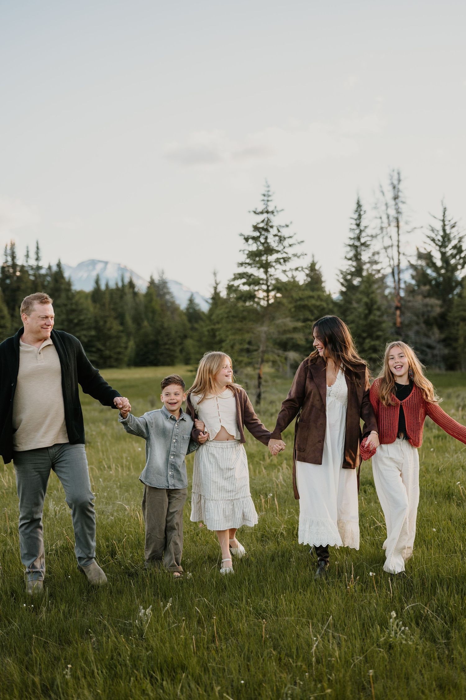 Happy family of five holding hands walking through a green meadow with pine trees and mountains in the background.