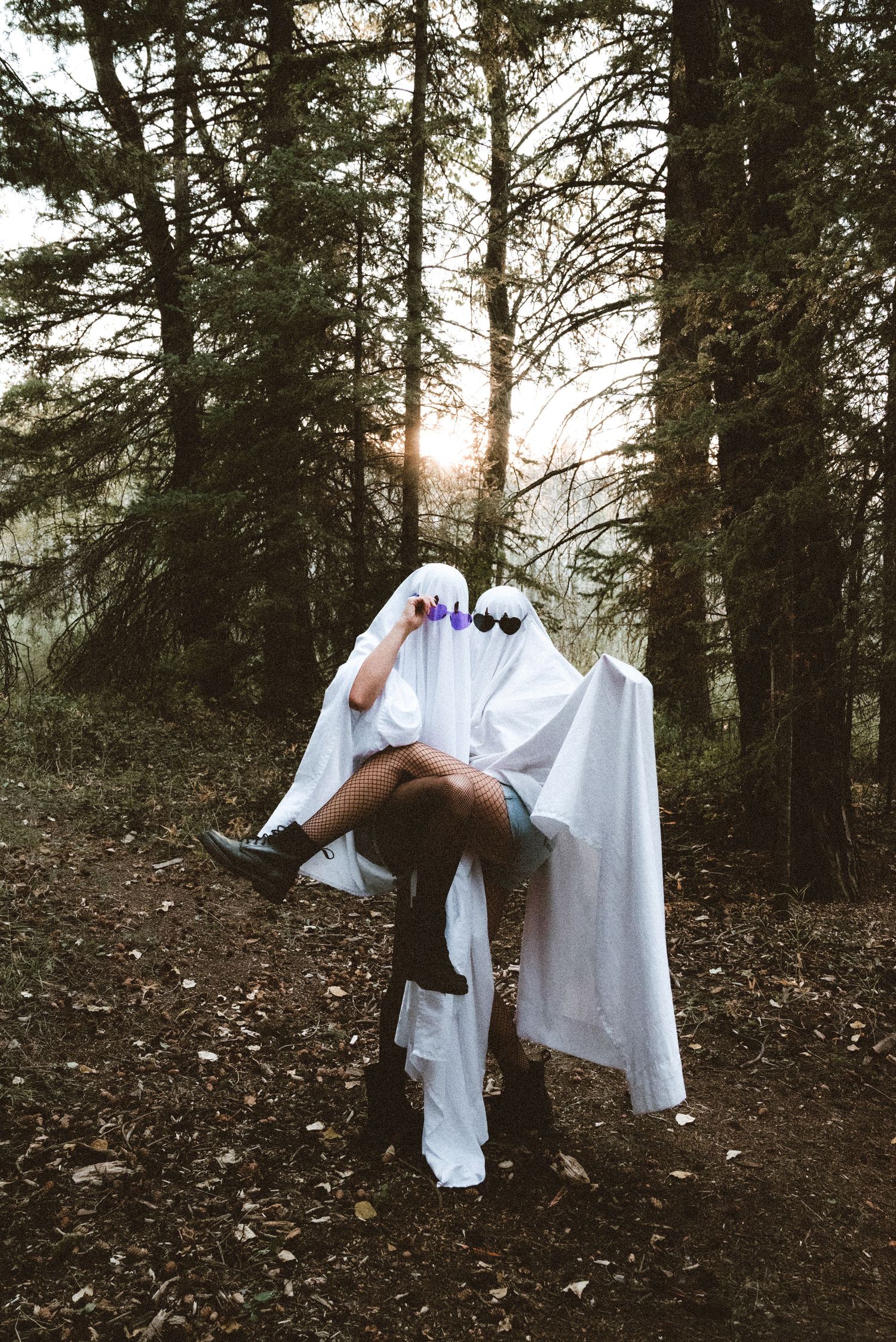 Two people in white ghost costumes embrace in a forest at sunset, one carrying the other.