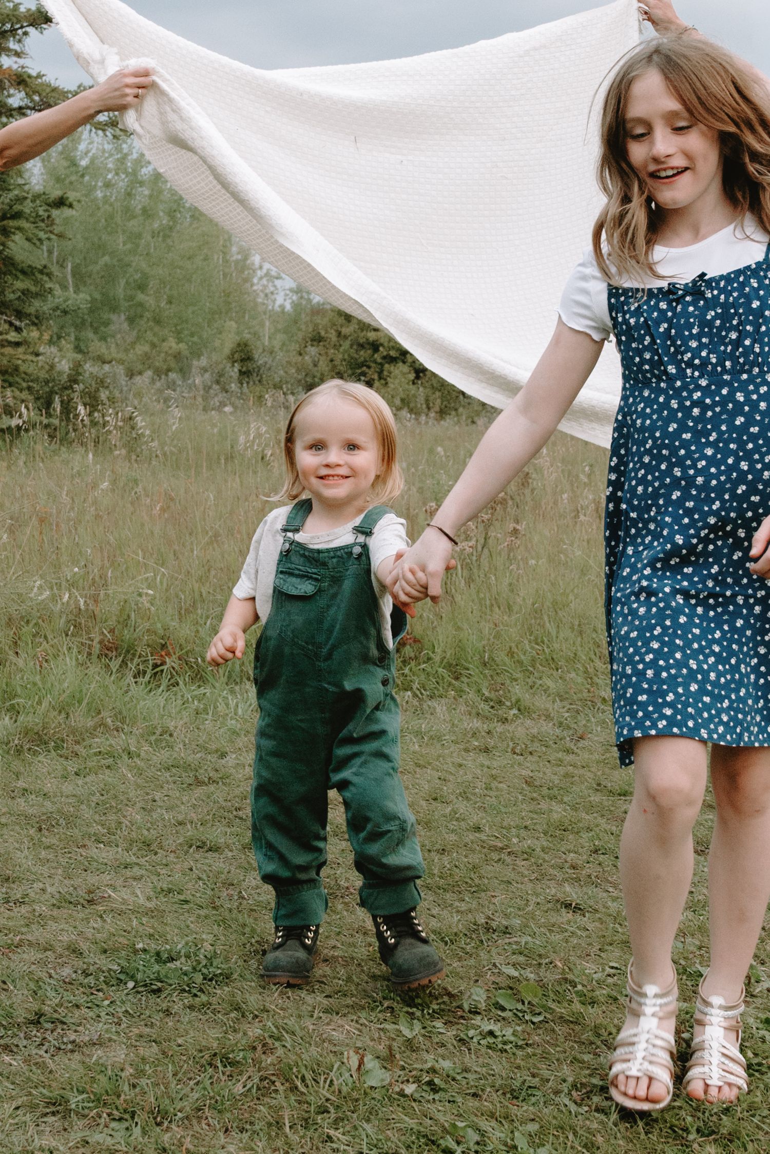 Two children holding hands in a grassy field, toddler in green overalls and girl in blue floral dress.