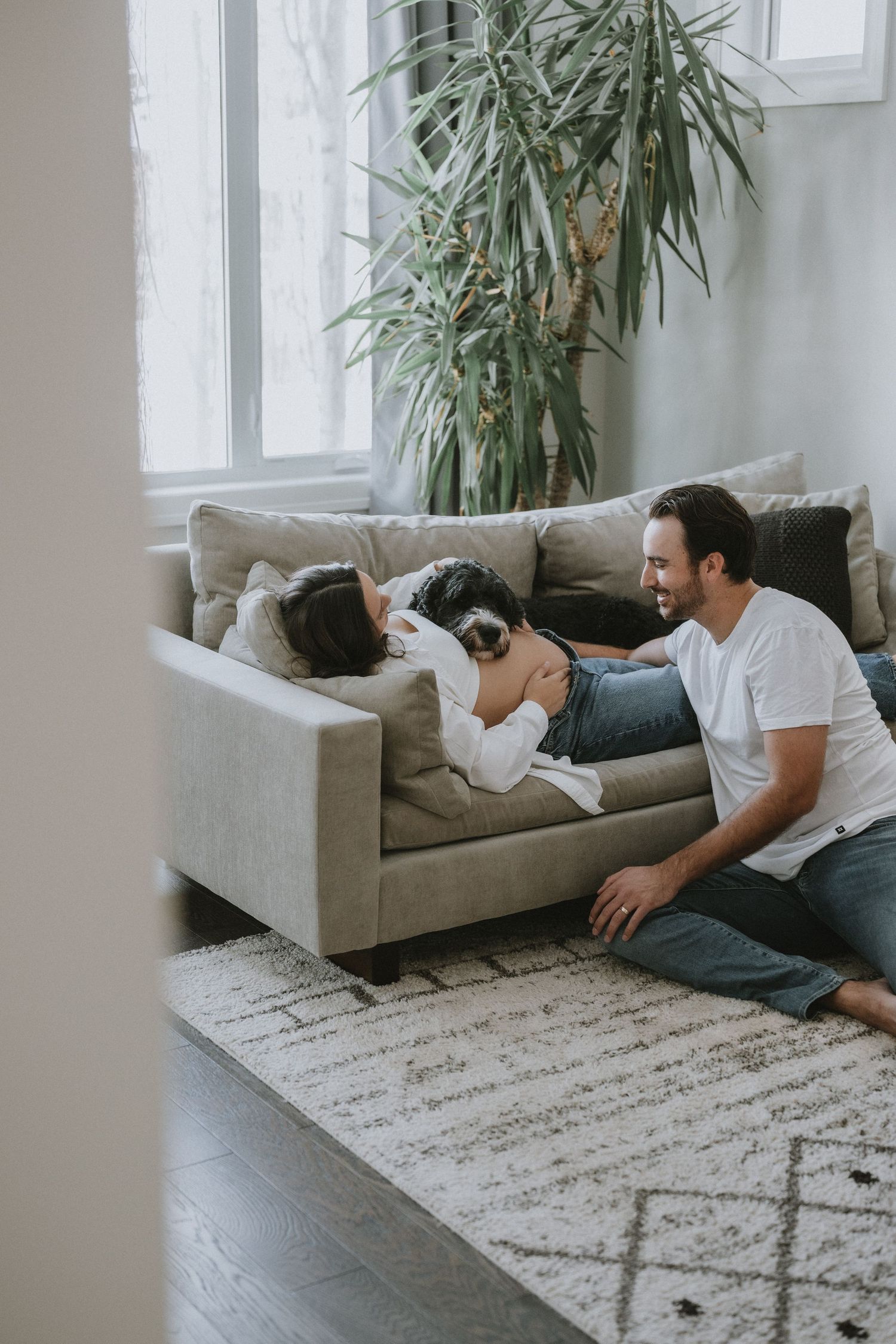 A man sits on the floor smiling at his pregnant partner lying on the couch with their dog.