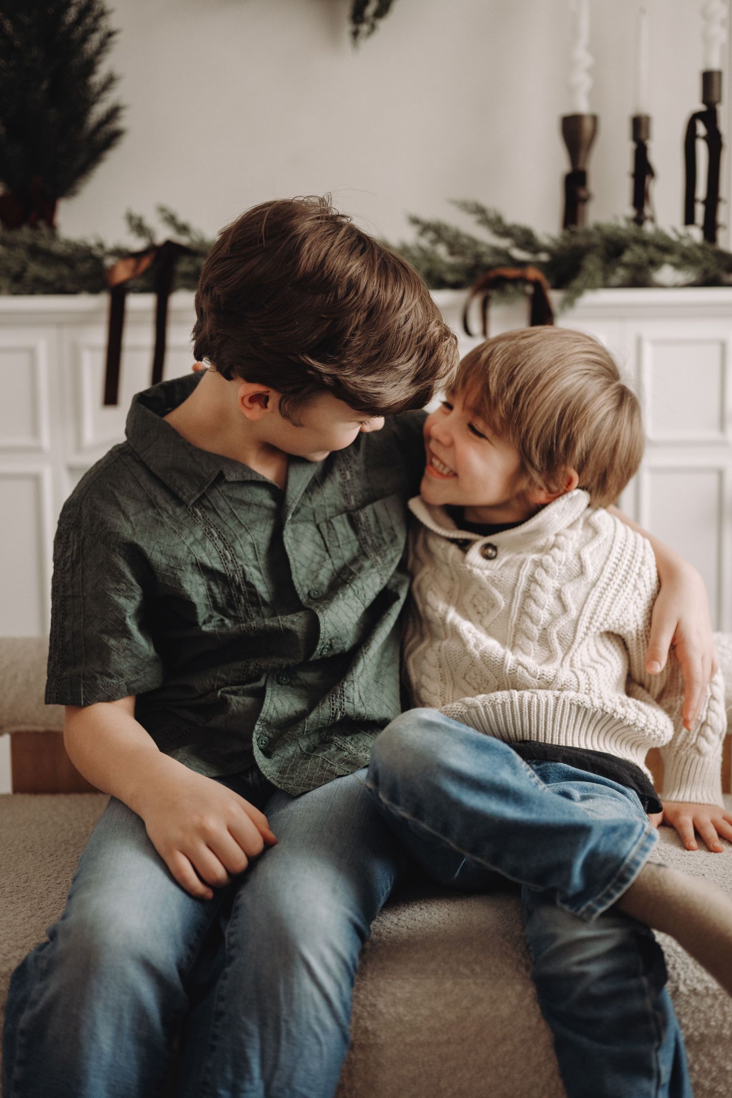 Two boys in festive sweaters share a sweet moment on stairs near holiday greenery decor.