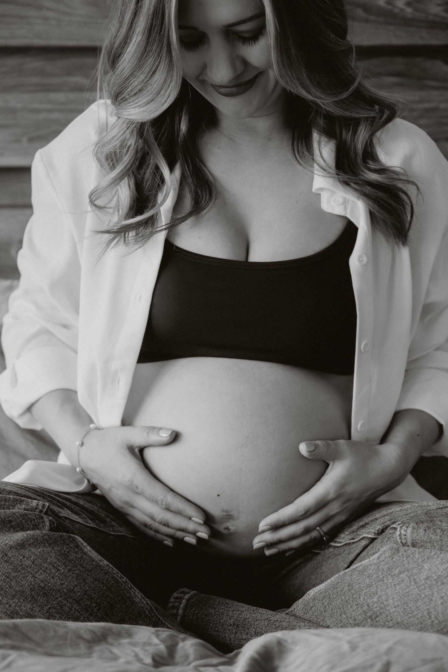 Black and white maternity photo of pregnant woman cradling baby bump, wearing open white shirt and black bralette.
