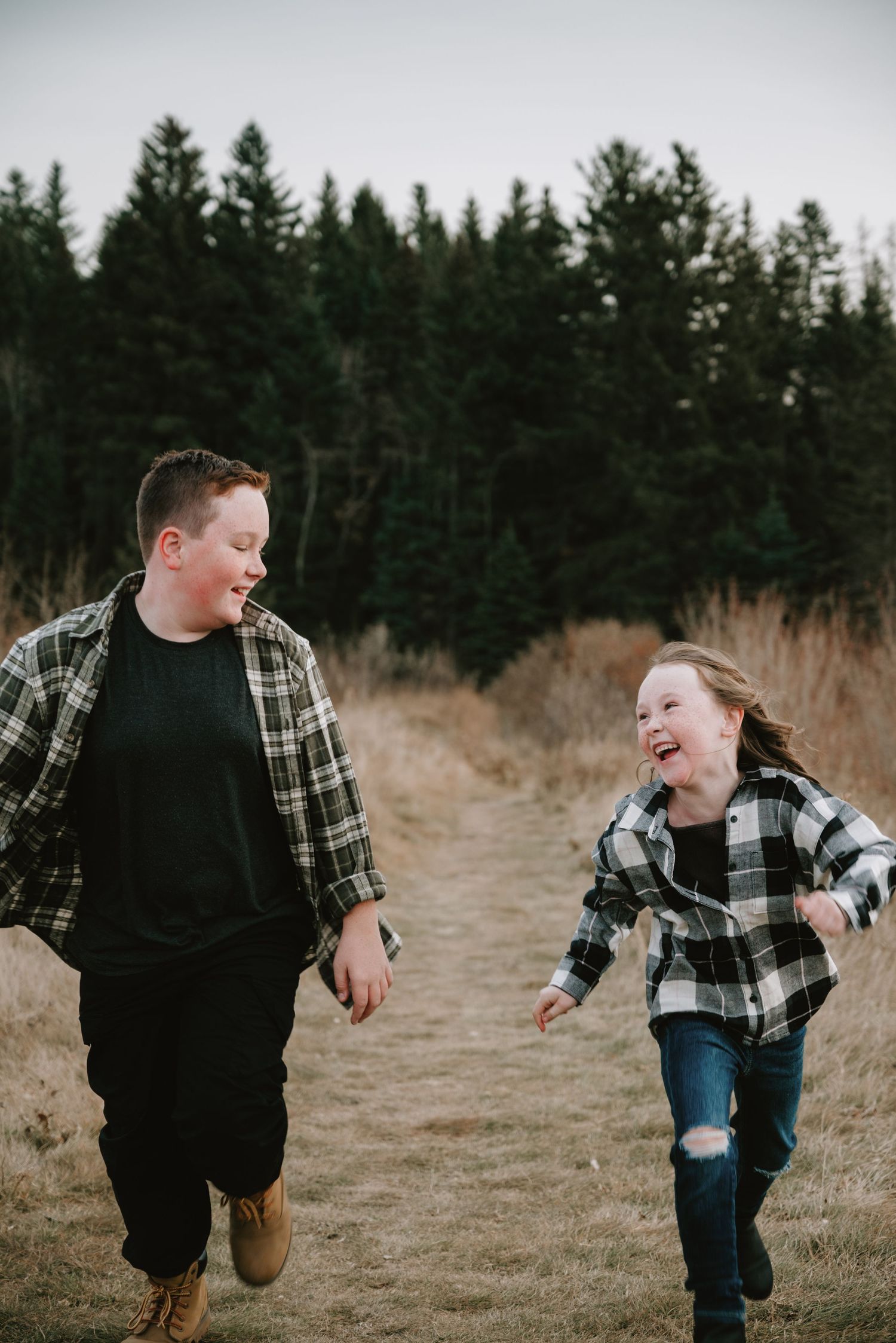 Two children in plaid shirts laugh and run together along a woodland path surrounded by tall evergreen trees.