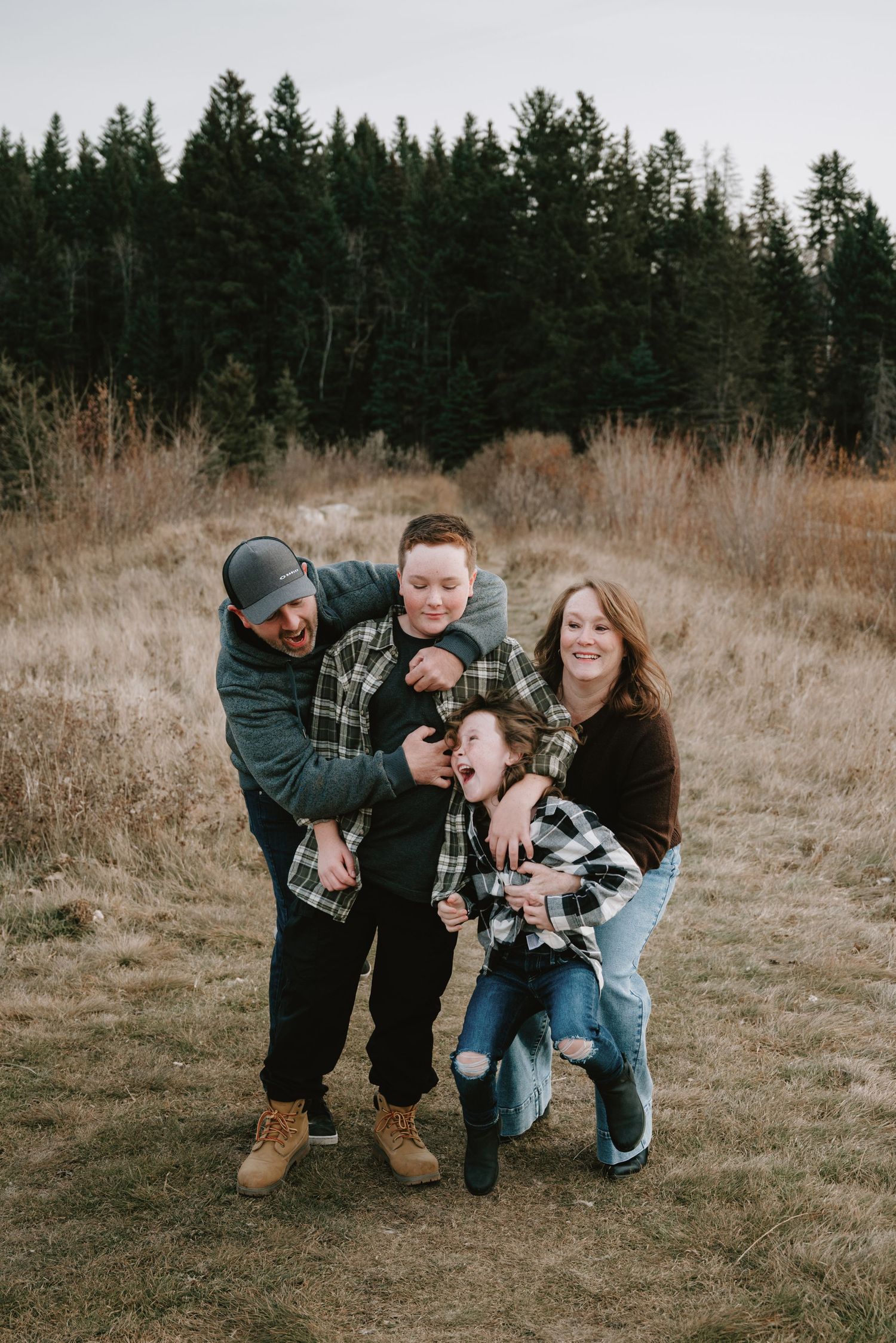 Family of four laughing and playing together in a rustic field with evergreen trees in the background during fall.