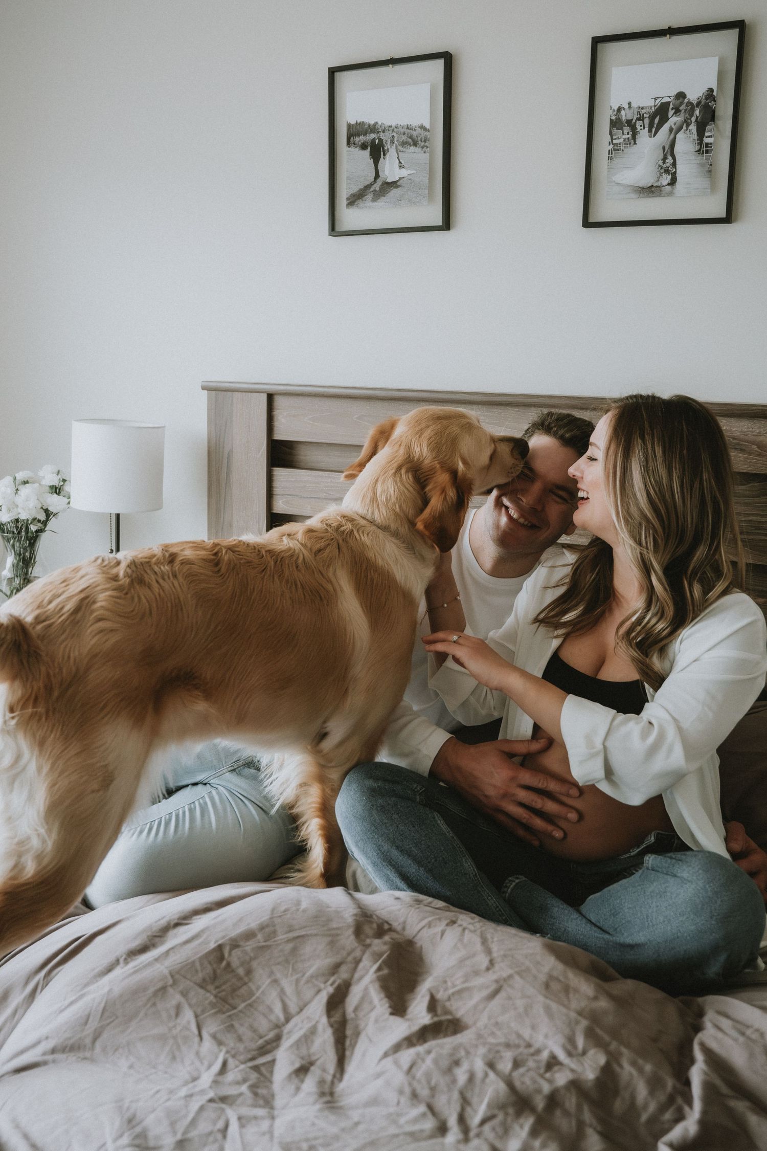 A couple sits on a bed smiling at each other while a golden retriever stands between them, with framed photos on the wall.