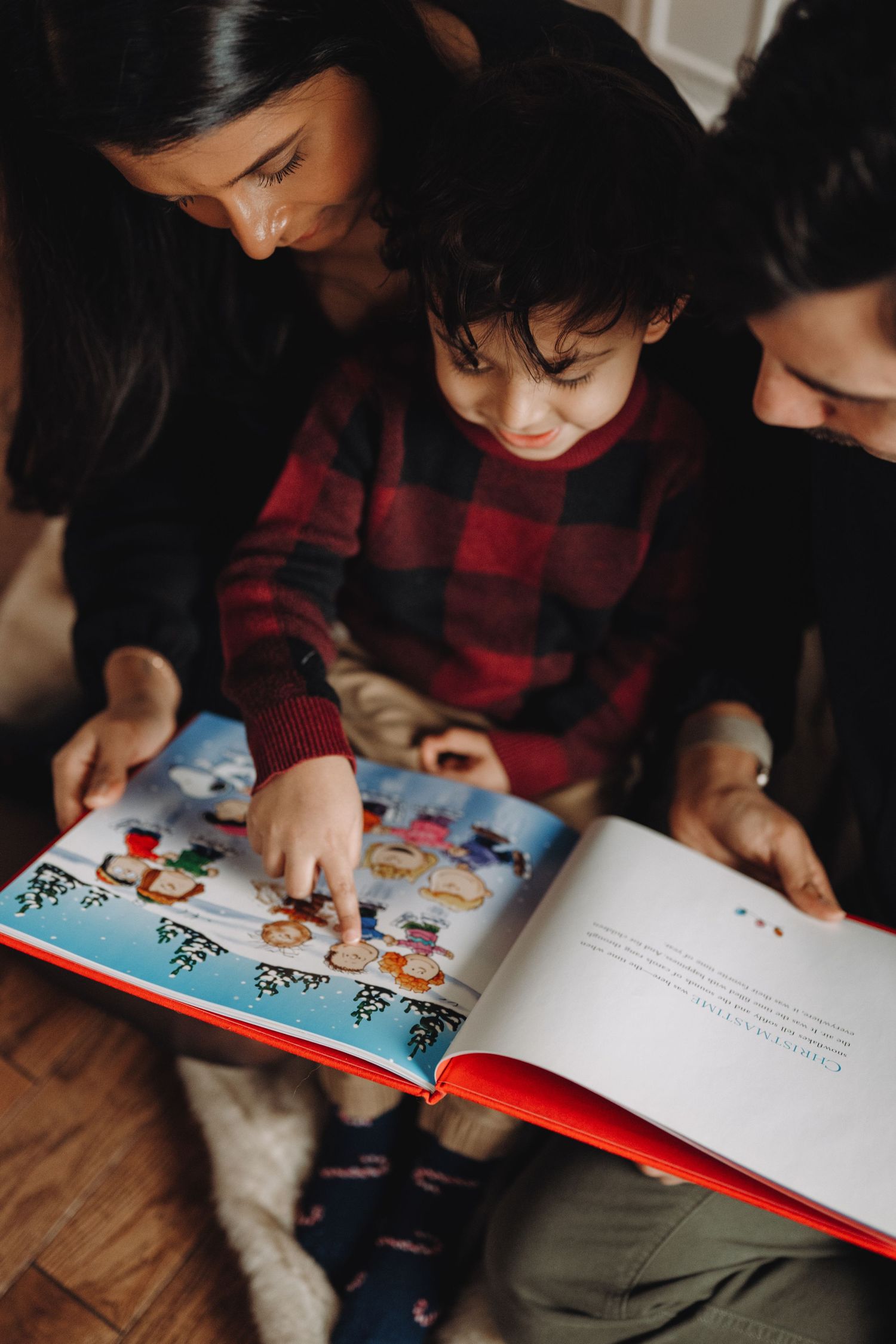Two women and a young child in plaid pajamas reading a colorful picture book together on the floor.
