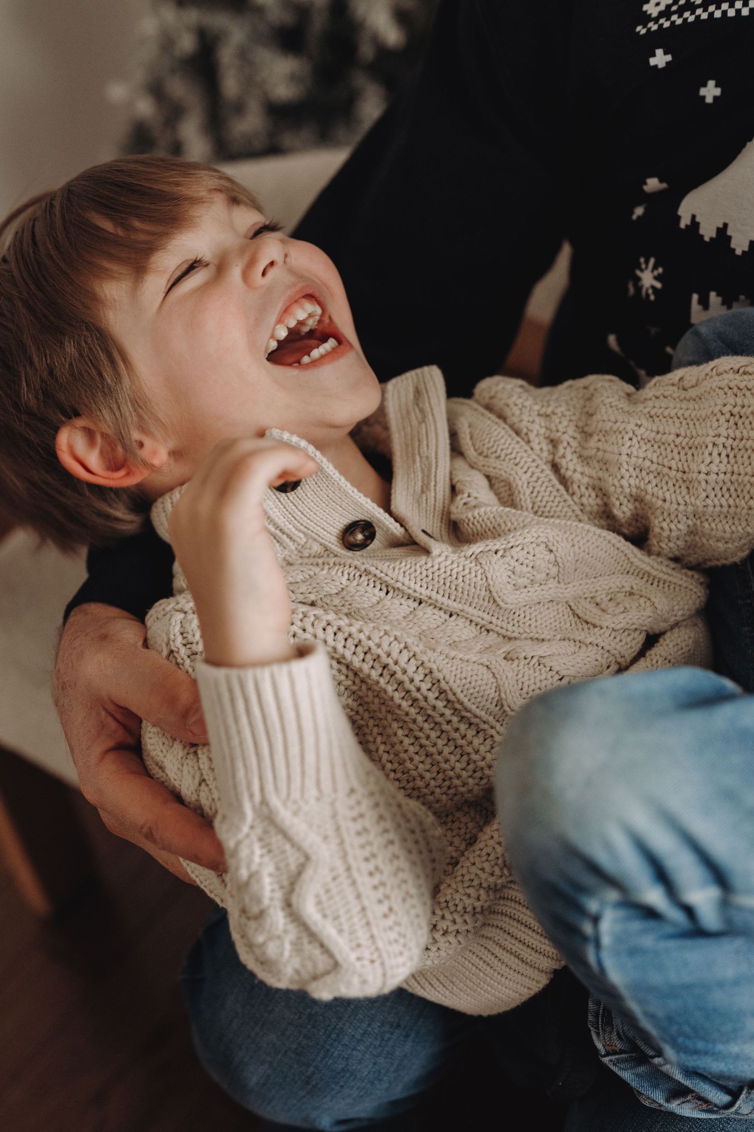 A joyful young boy in a cream knit sweater laughs while being held by an adult near a Christmas tree.