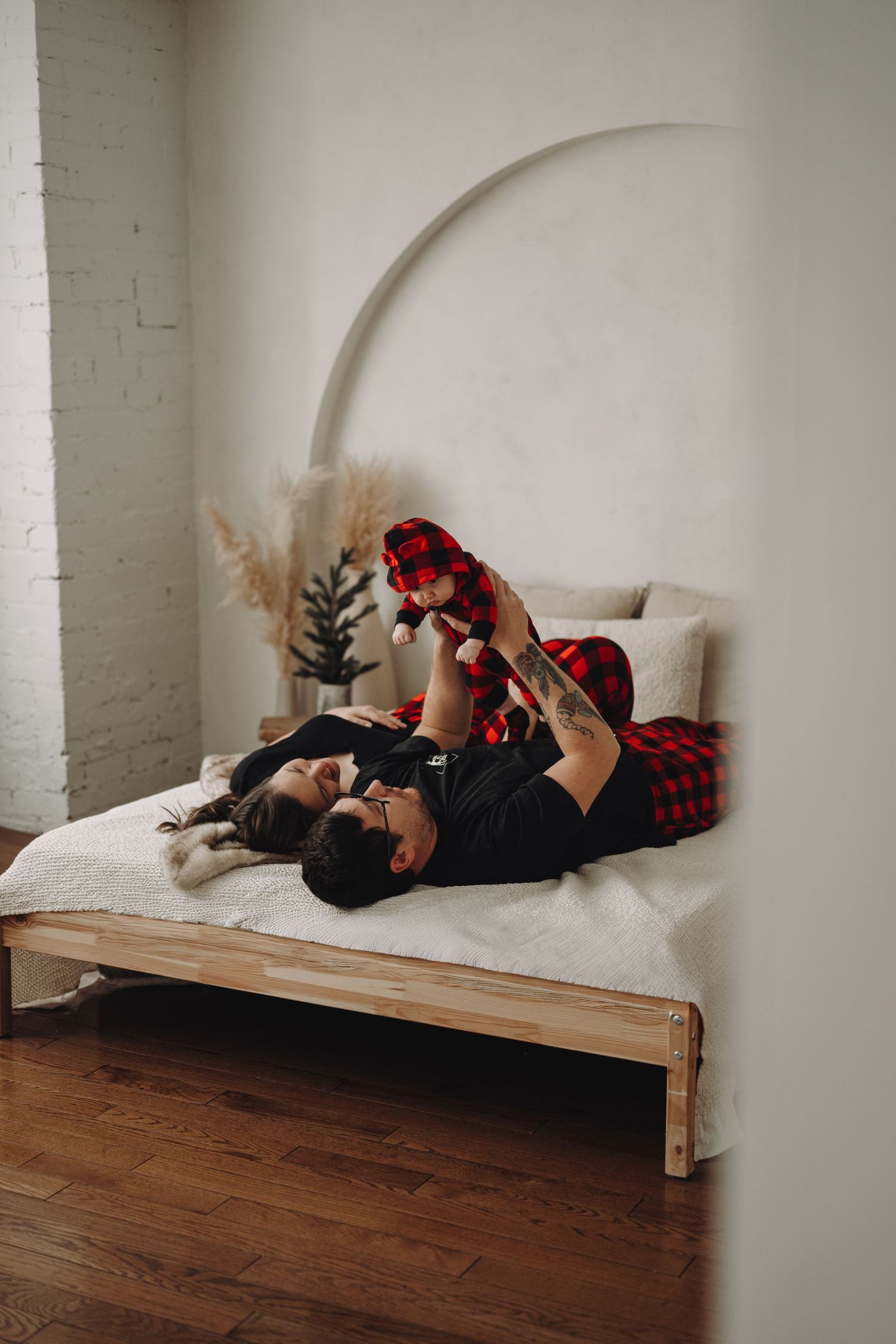 Couple lying on a wooden bed in matching red plaid pajamas, playing with their baby in a modern white bedroom.