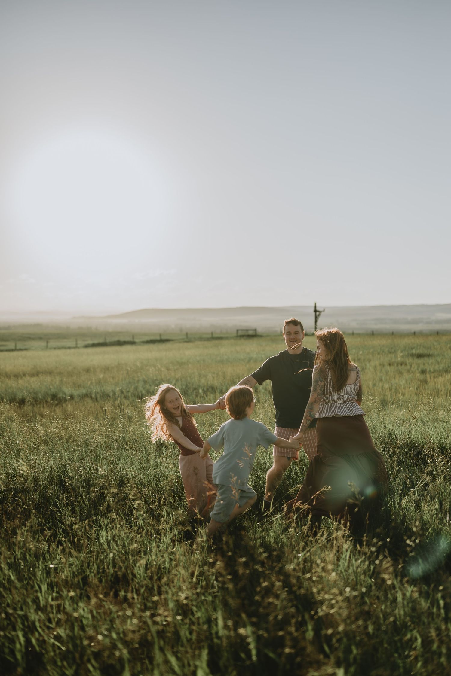Family of four holding hands in a circle, playing in a sunny green field at golden hour.
